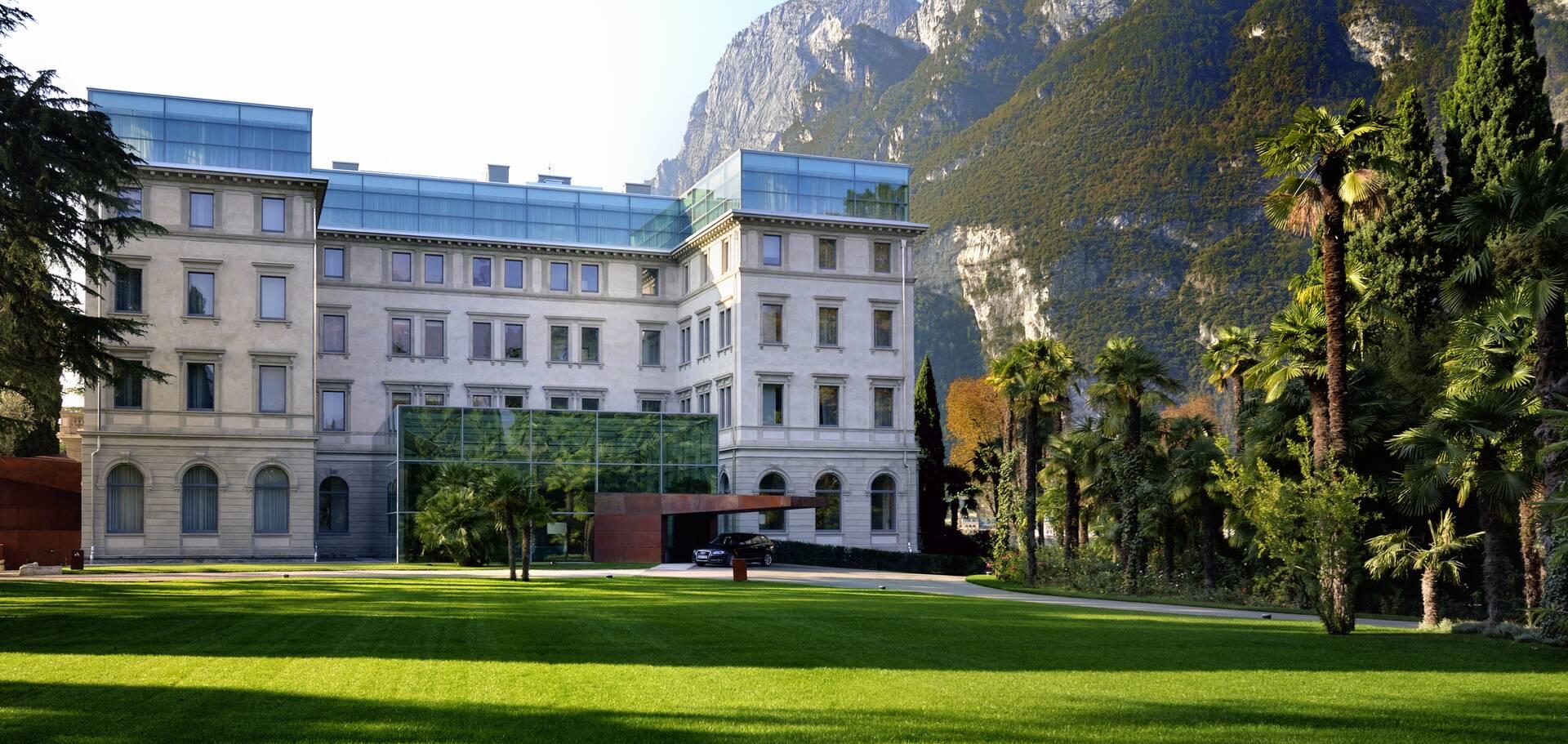 The hotel's stone facade seen across a wide lawn, with a glass-topped wing, palm trees, and a steep mountain behind.