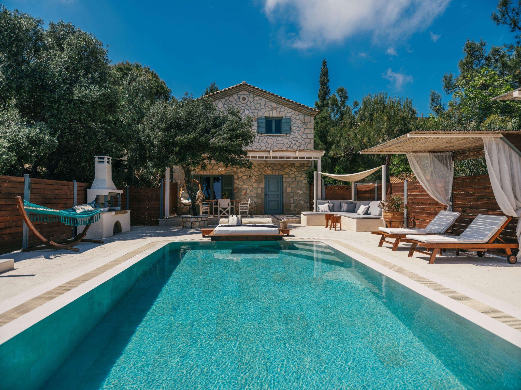 a two-storey stone villa with blue shutters seen across a swimming pool, with a teal hammock, a stone barbecue and a shaded outdoor sofa on the terrace in front of it
