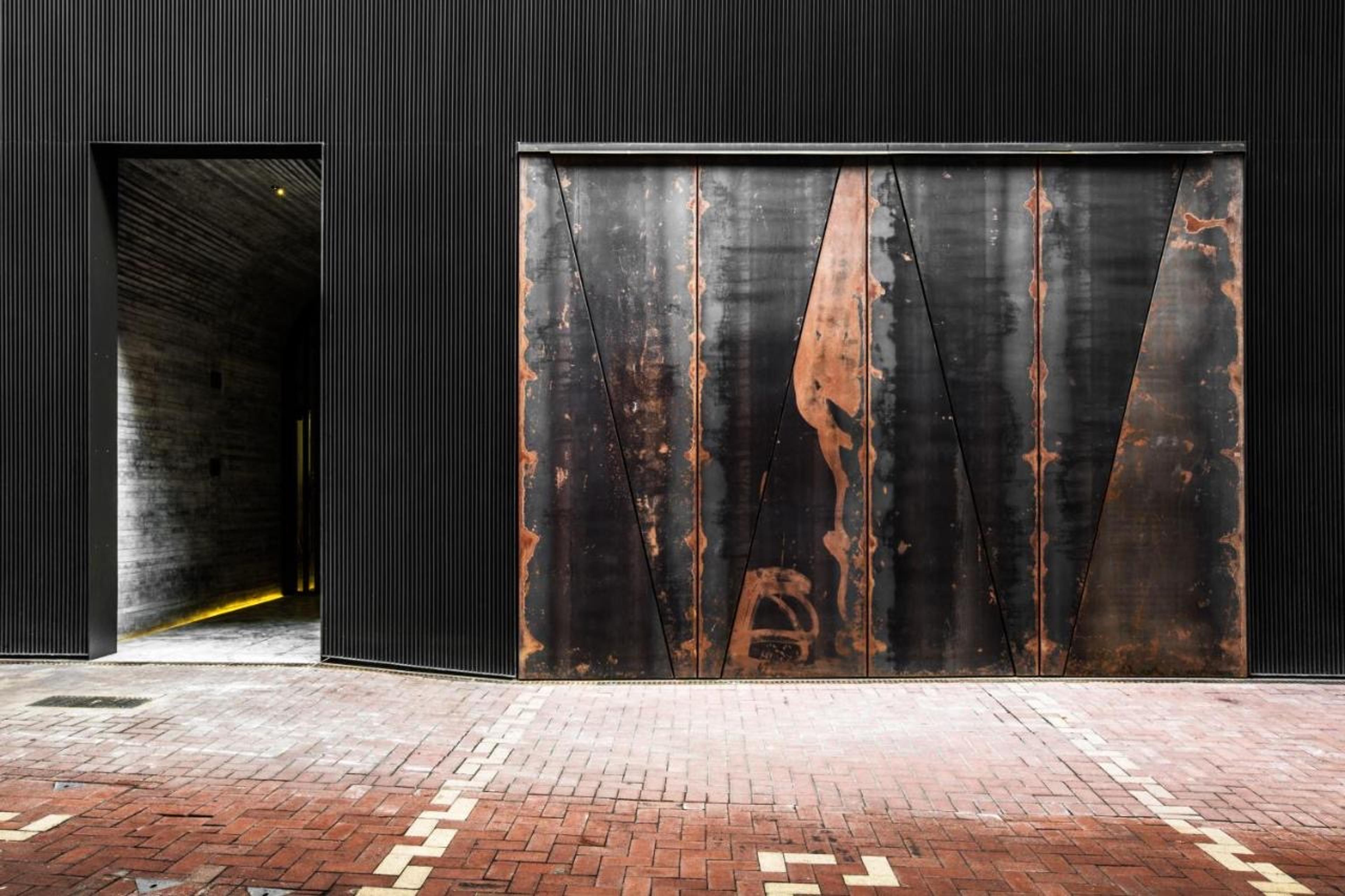 A building's dark corrugated facade at street level, with a rusted steel door beside an open doorway lit from within, set above a brick pavement.