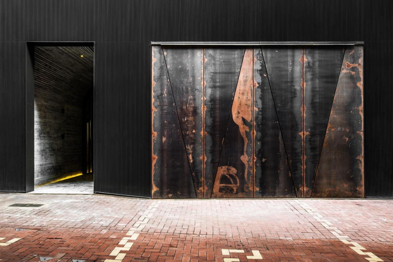 A building's dark corrugated facade at street level, with a rusted steel door beside an open doorway lit from within, set above a brick pavement.