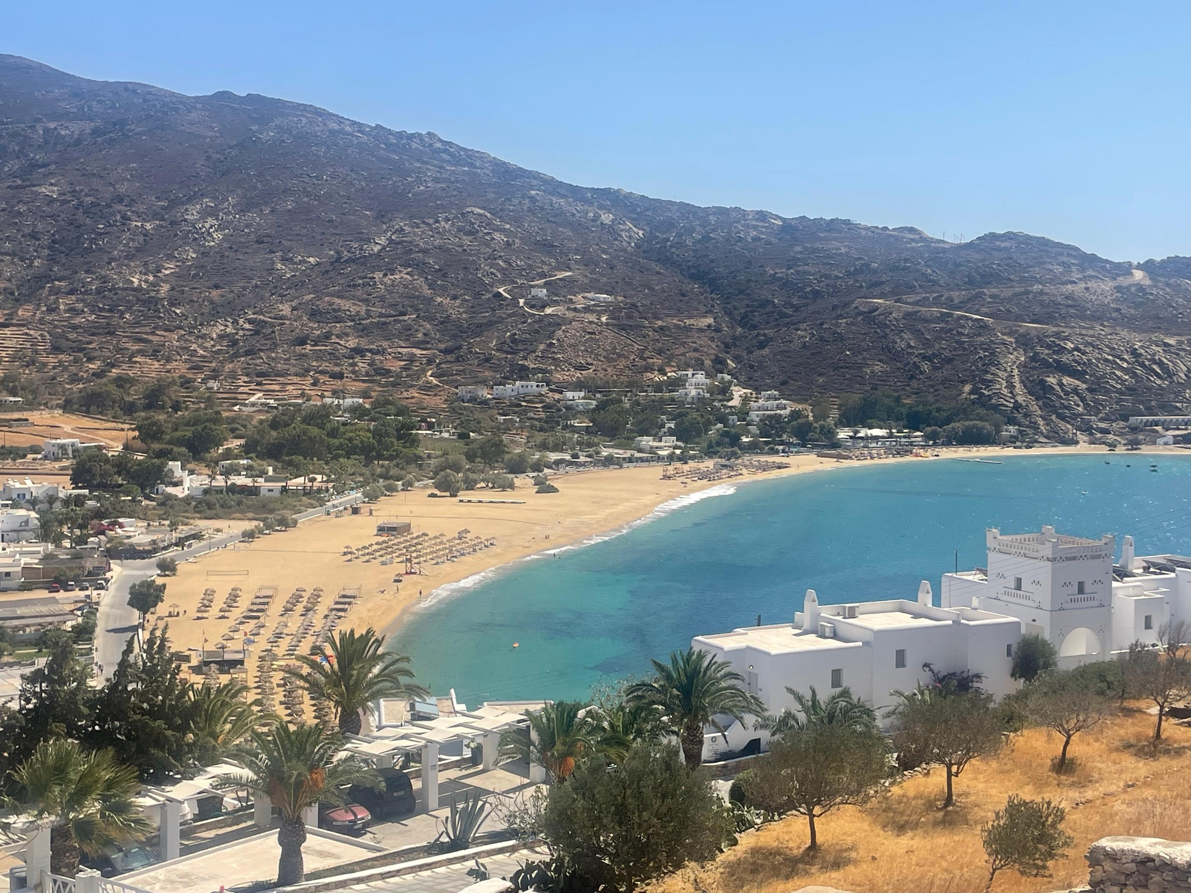 Mylopotas bay on Ios from the hillside above, a curve of sand and turquoise water under bare brown hills, white houses and palms in the foreground