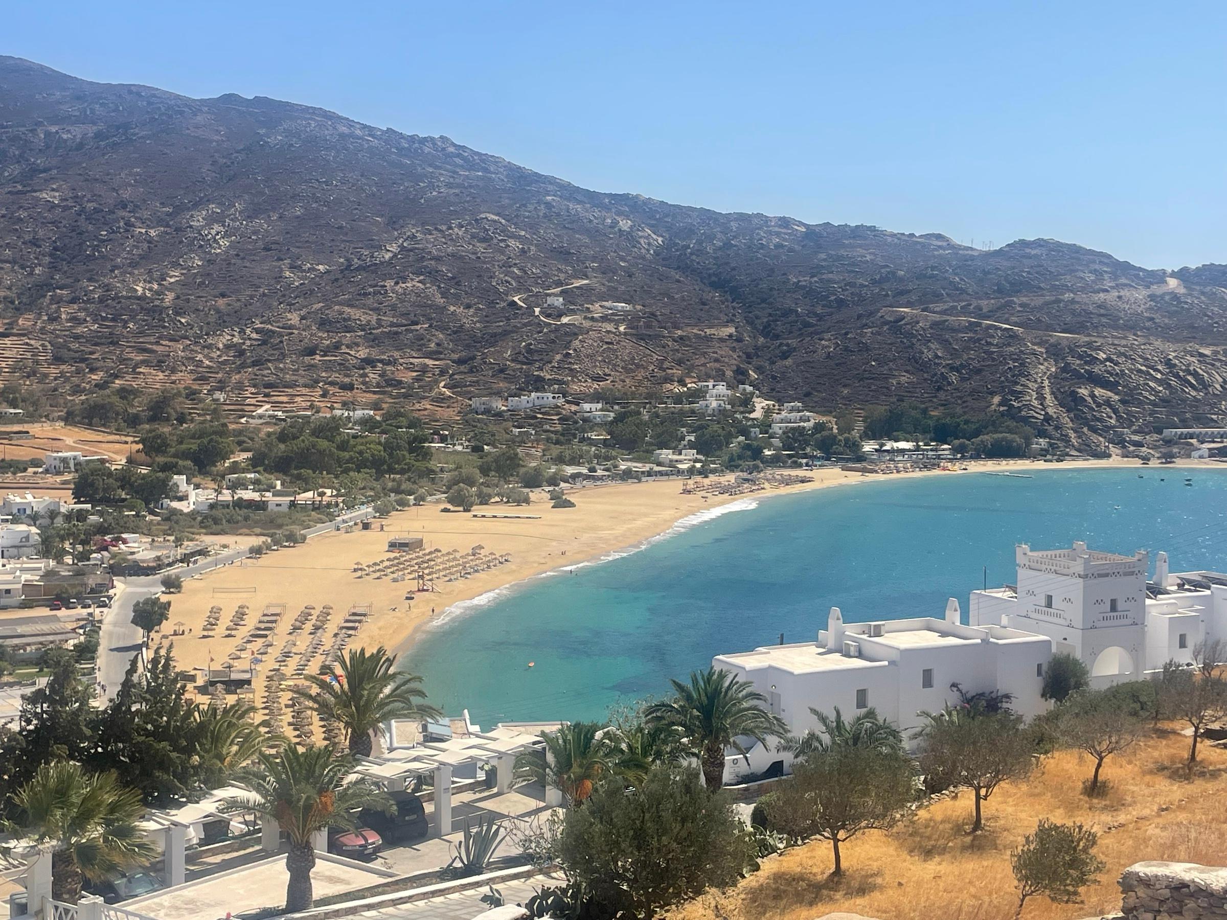 Mylopotas bay on Ios from the hillside above, a curve of sand and turquoise water under bare brown hills, white houses and palms in the foreground