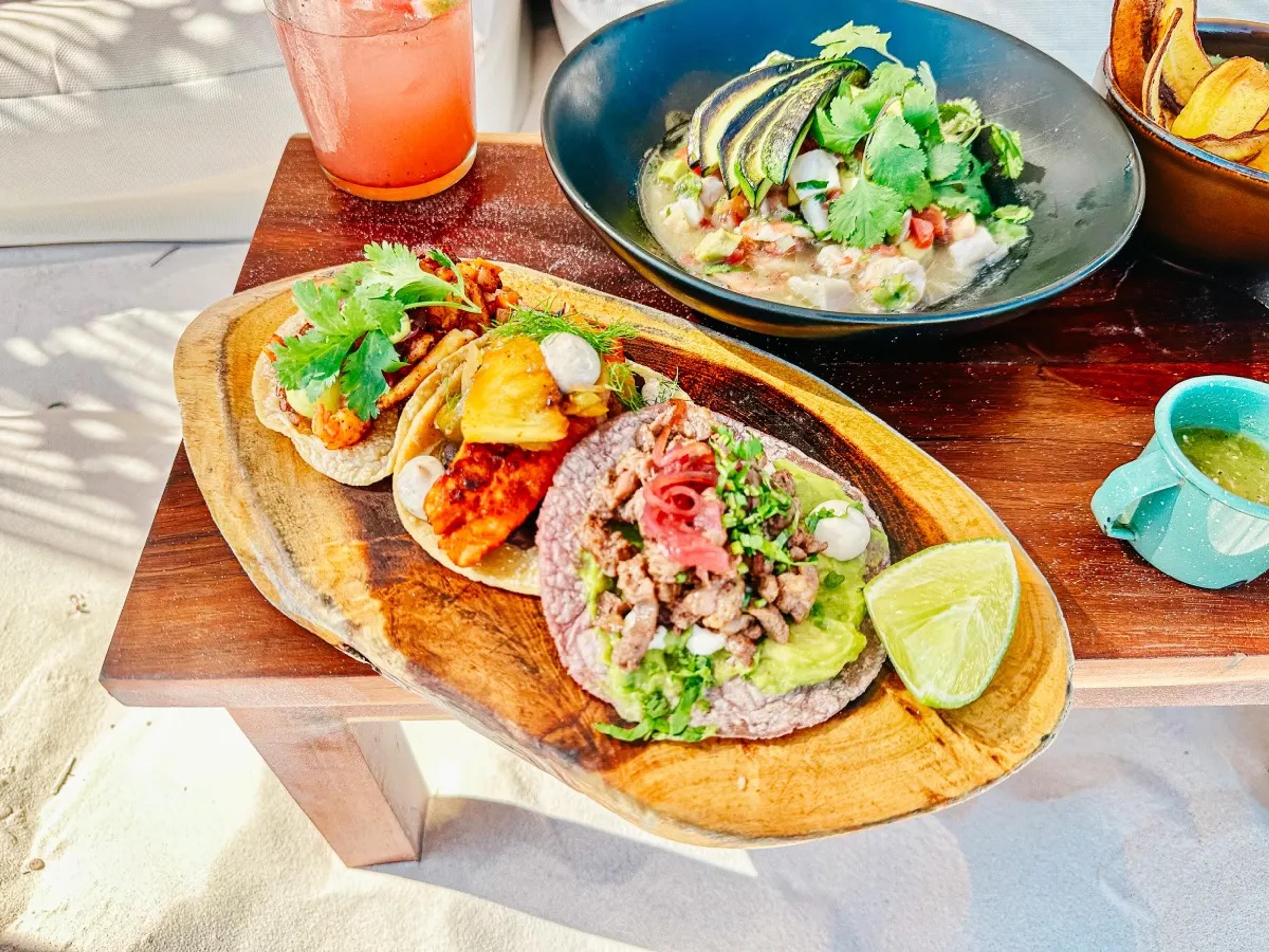 Tacos and lime on a wooden board on the sand, a drink and a bowl of guacamole beside them
