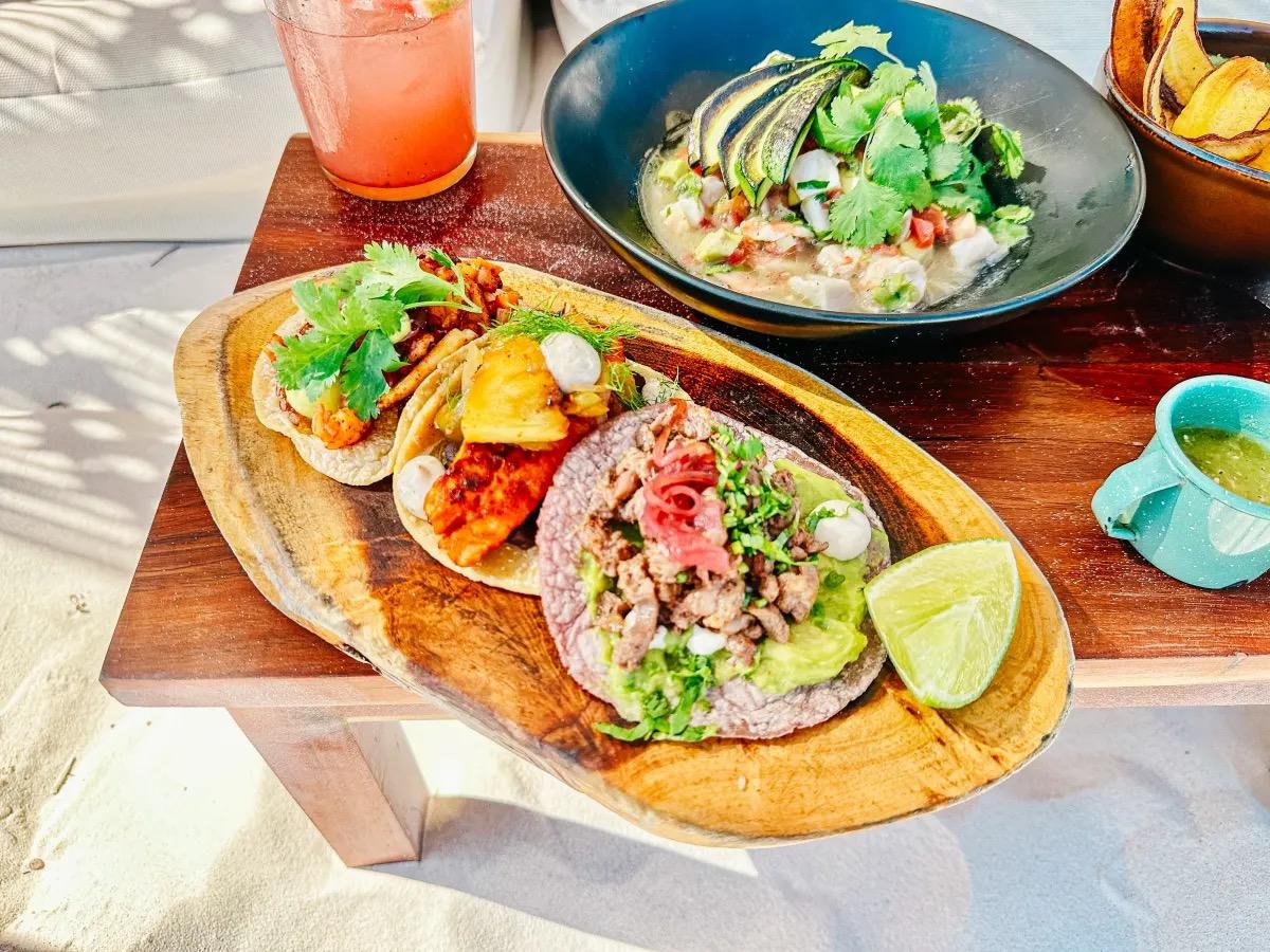 Tacos and lime on a wooden board on the sand, a drink and a bowl of guacamole beside them