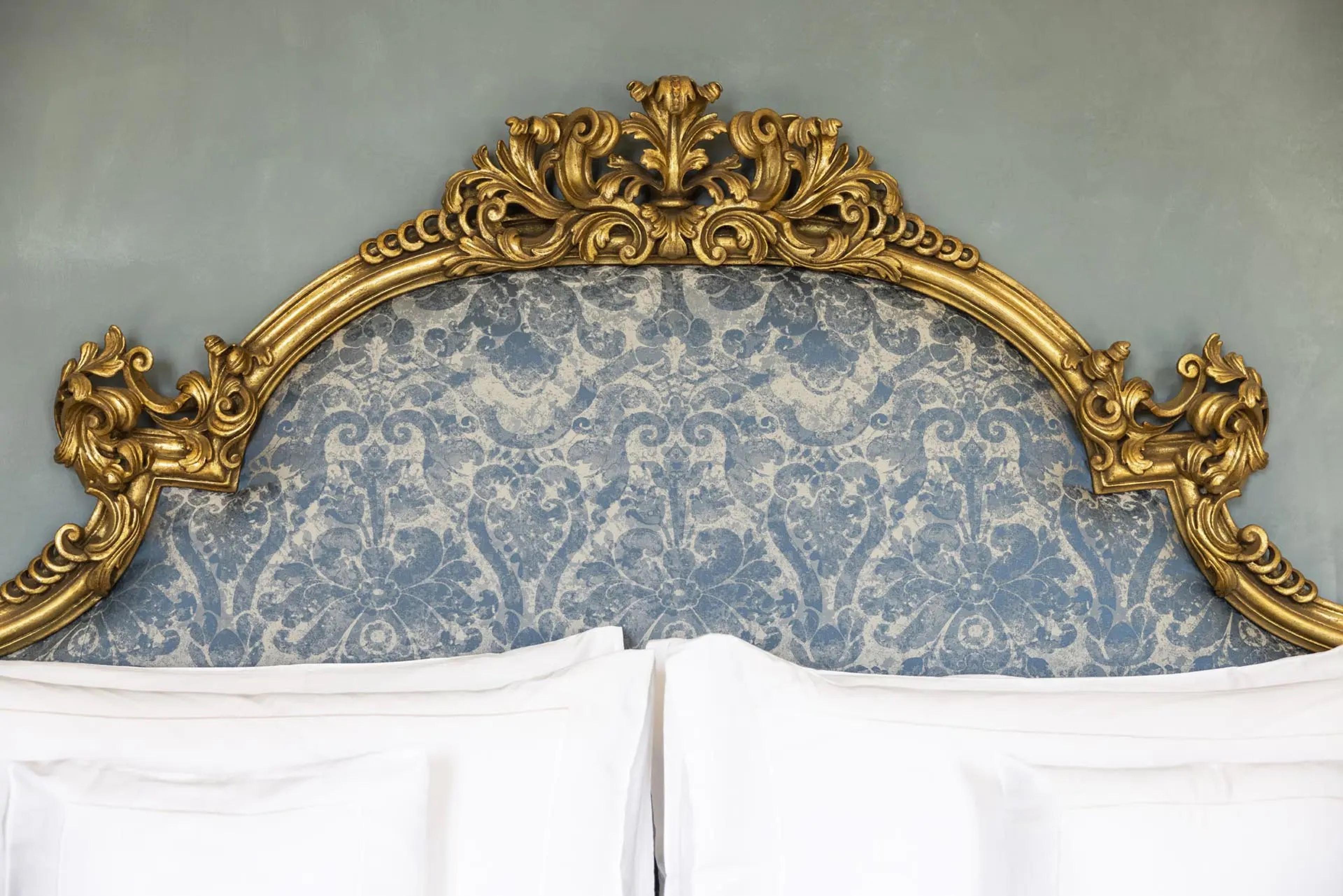 A gilded scalloped headboard with faded blue and cream damask fabric, above two pillows on an unmade bed.