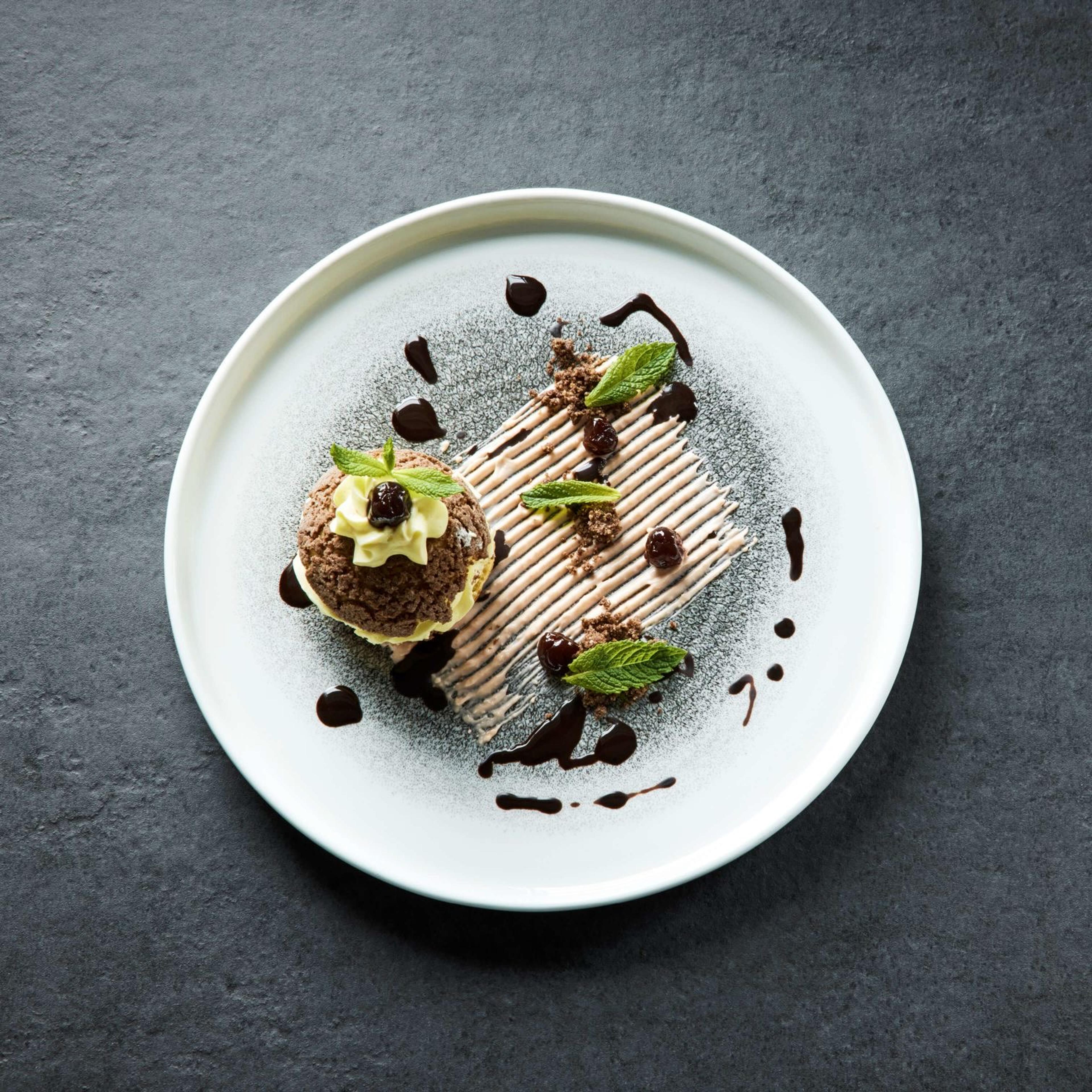 A plated dessert of chocolate sponge, cream and cherries with mint leaves and a chocolate drizzle, dusted with icing sugar on a dark surface.