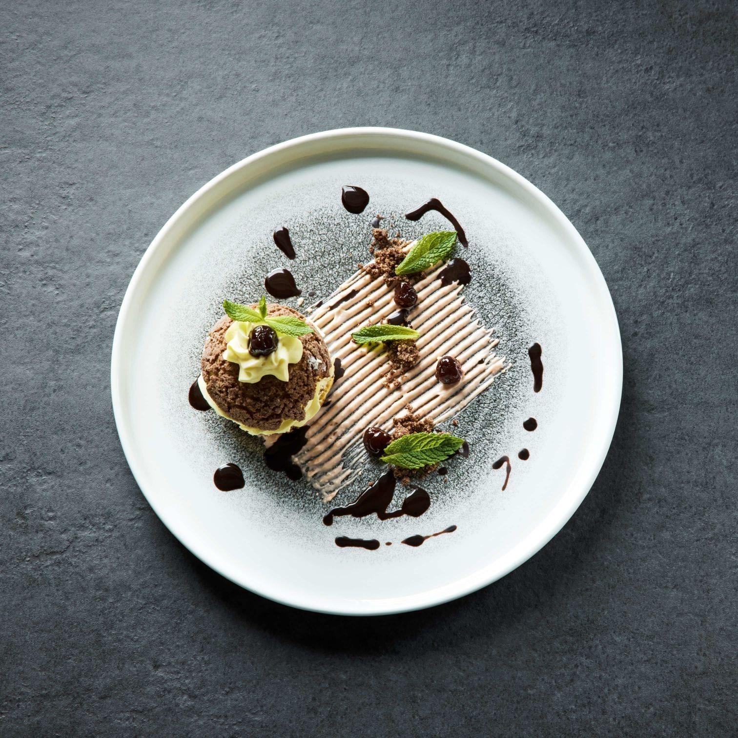 A plated dessert of chocolate sponge, cream and cherries with mint leaves and a chocolate drizzle, dusted with icing sugar on a dark surface.