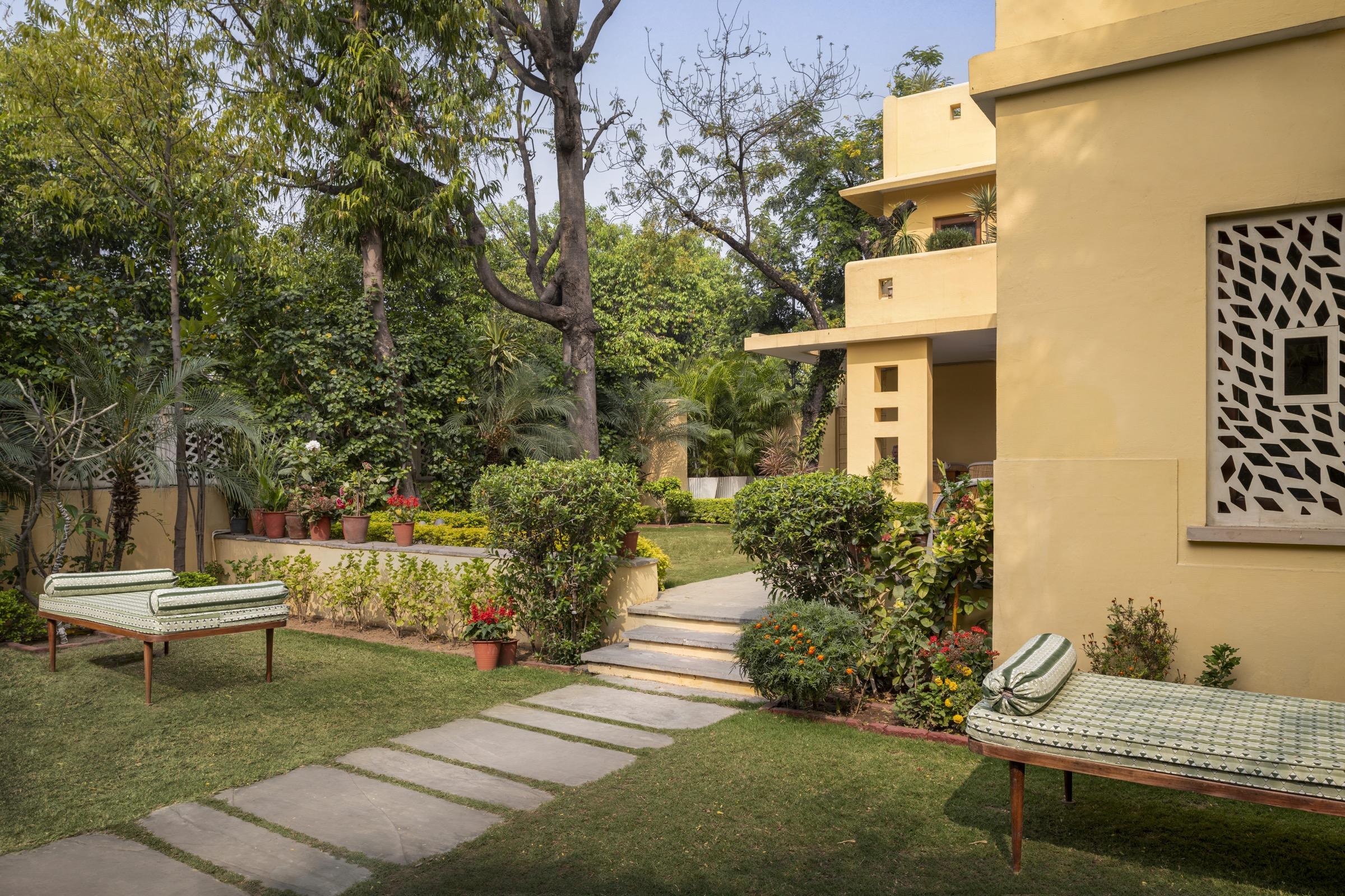 Two daybeds on a lawn beside a path to a yellow building with a carved stone screen window, pine and palm trees behind.