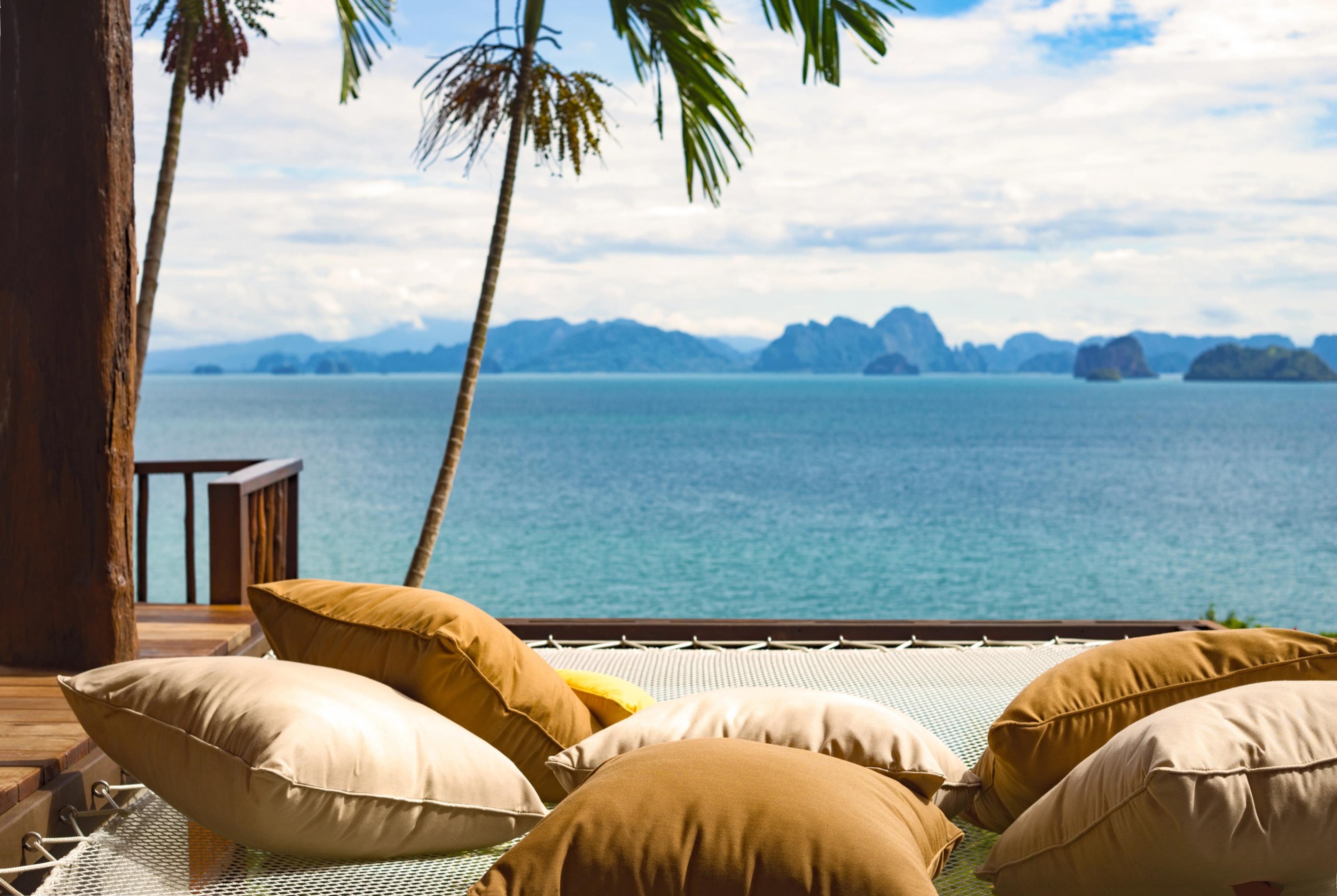 A net hammock strung with cushions above the sea, framed by palm trees and a view of distant islands under a partly cloudy sky.