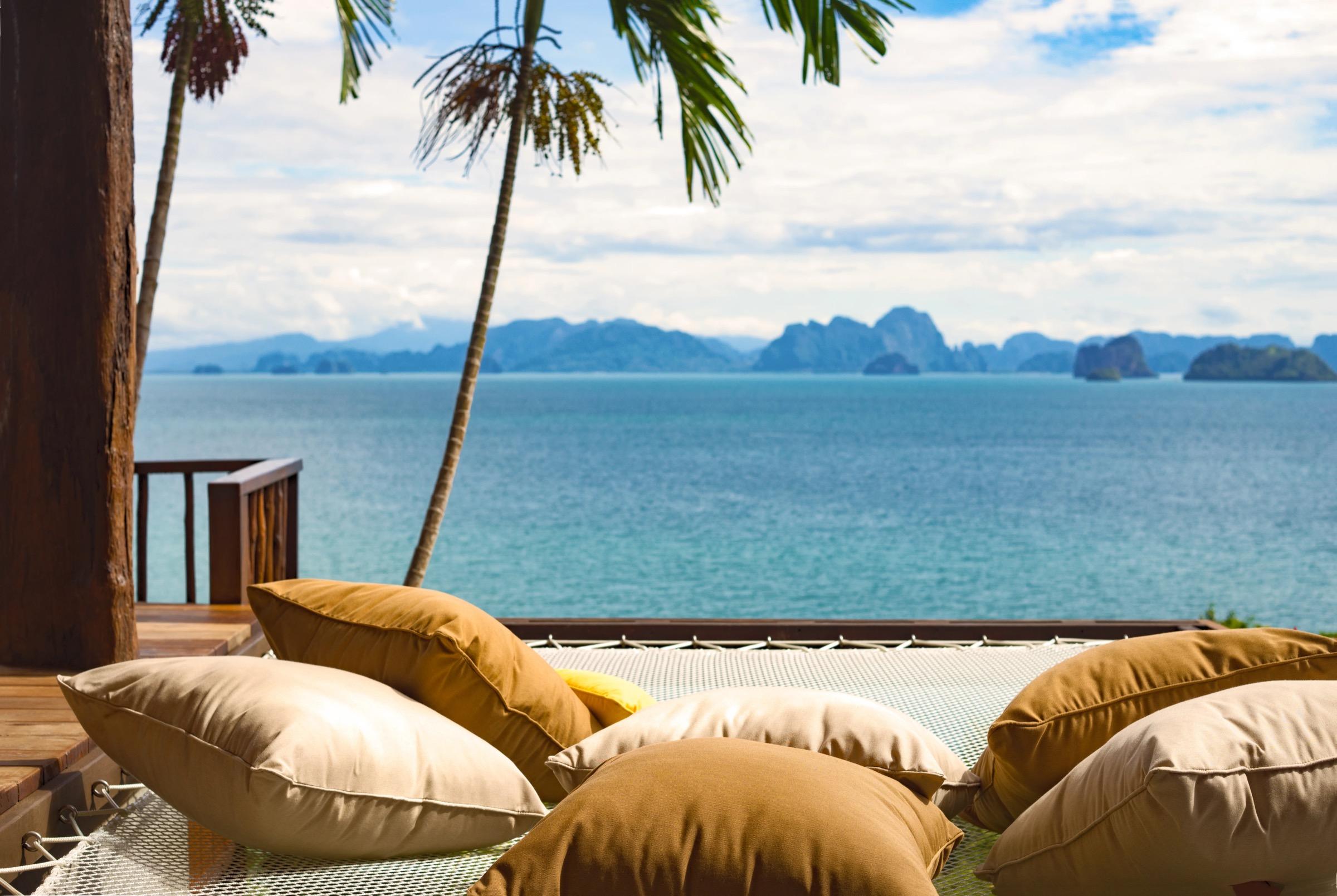 A net hammock strung with cushions above the sea, framed by palm trees and a view of distant islands under a partly cloudy sky.