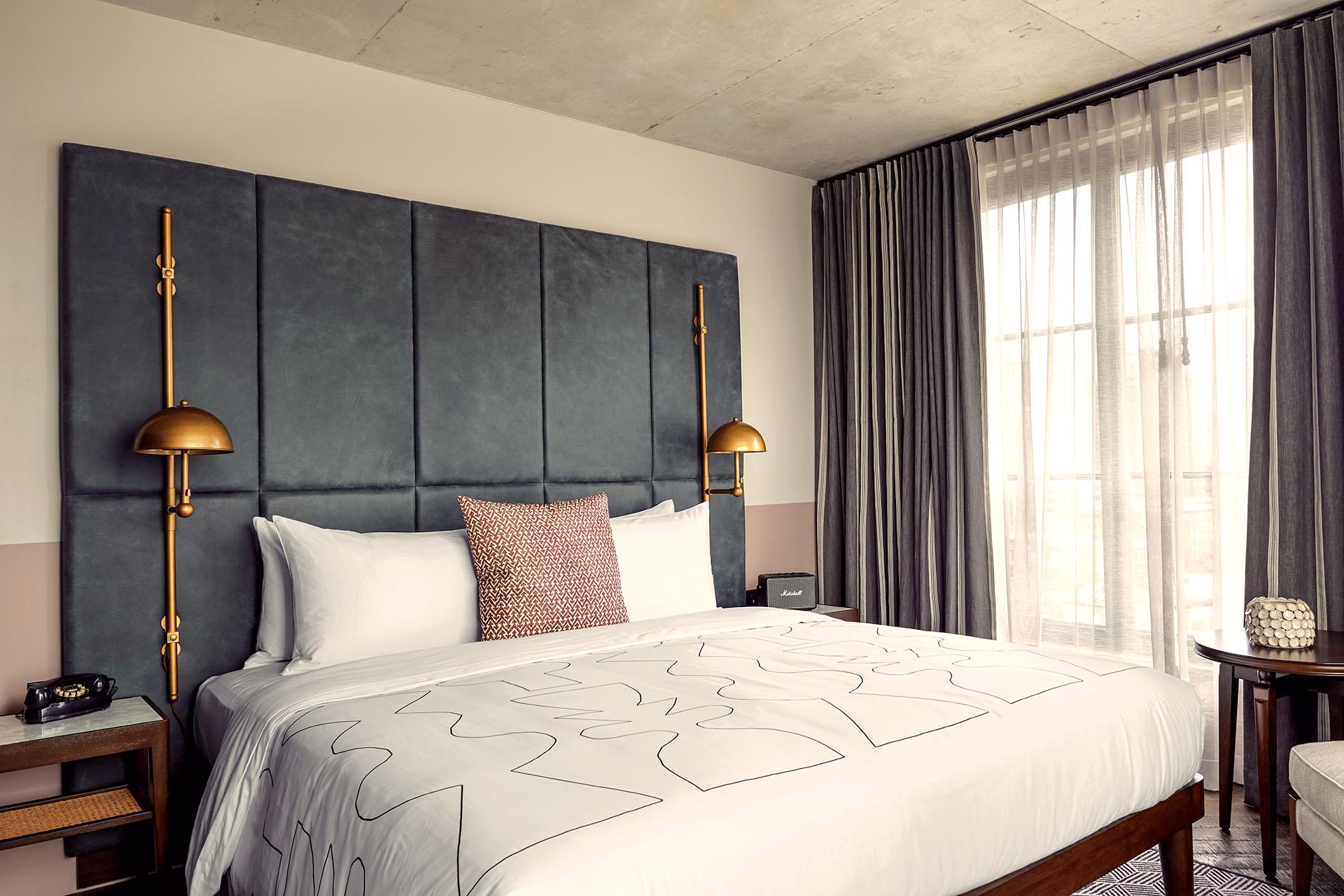 A king bed with a charcoal panelled headboard and brass reading lamps, facing curtained windows with a round side table and armchair.