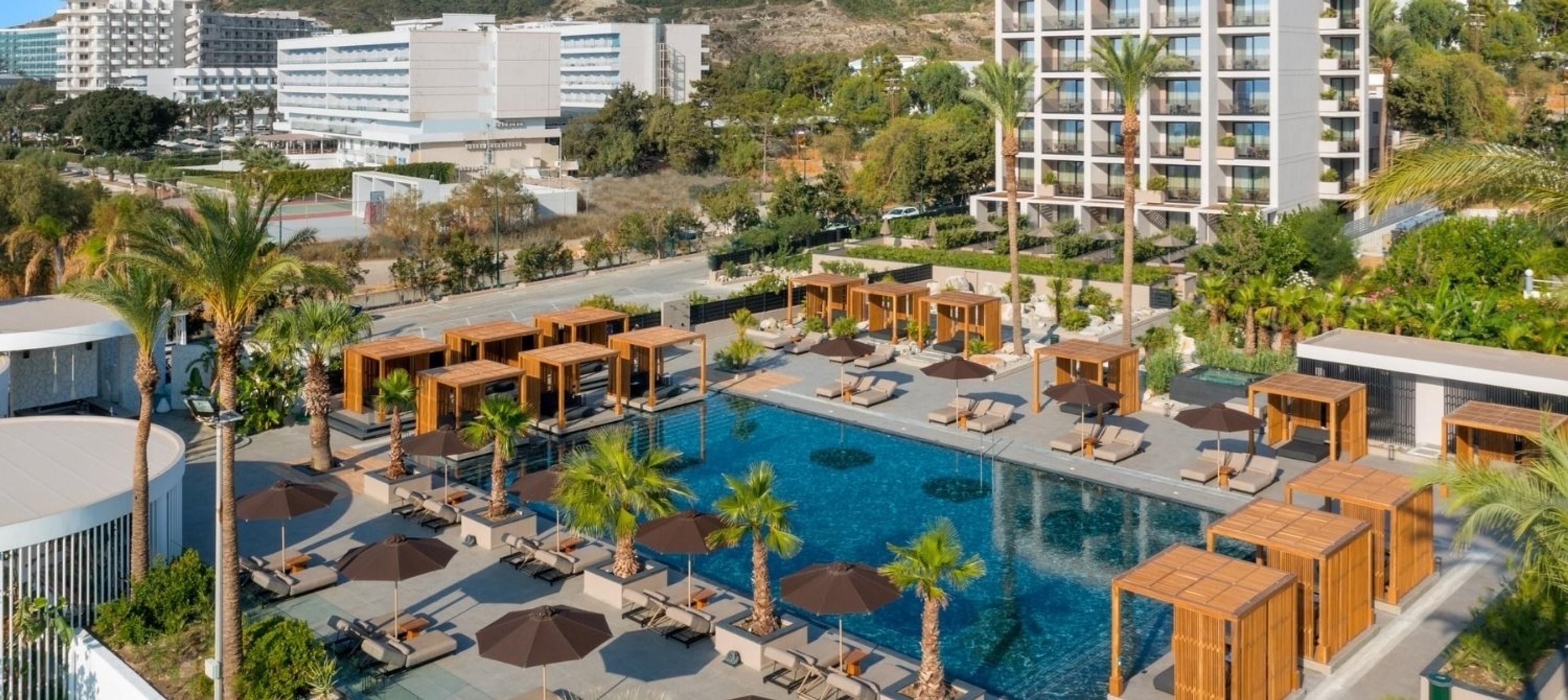 An aerial view of the pool, wooden cabanas and sun loungers, with the hotel buildings and hills beyond.