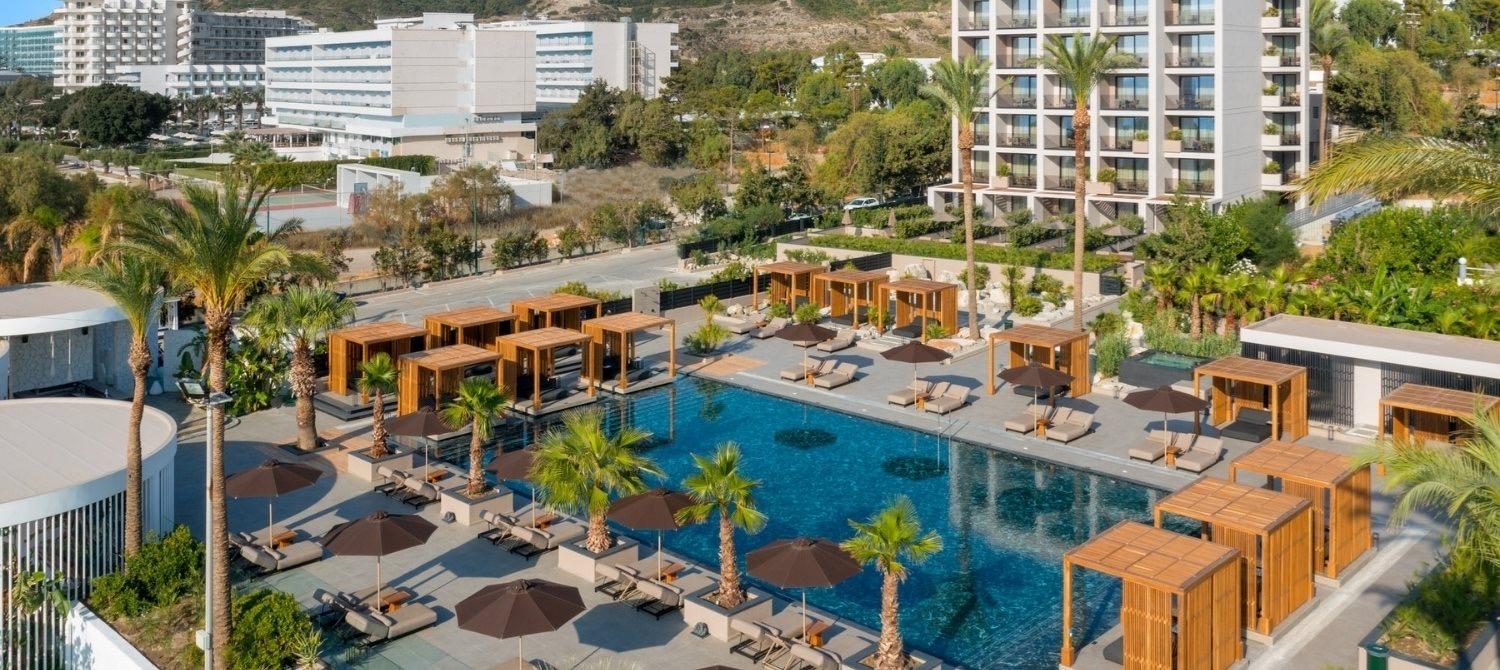 An aerial view of the pool, wooden cabanas and sun loungers, with the hotel buildings and hills beyond.