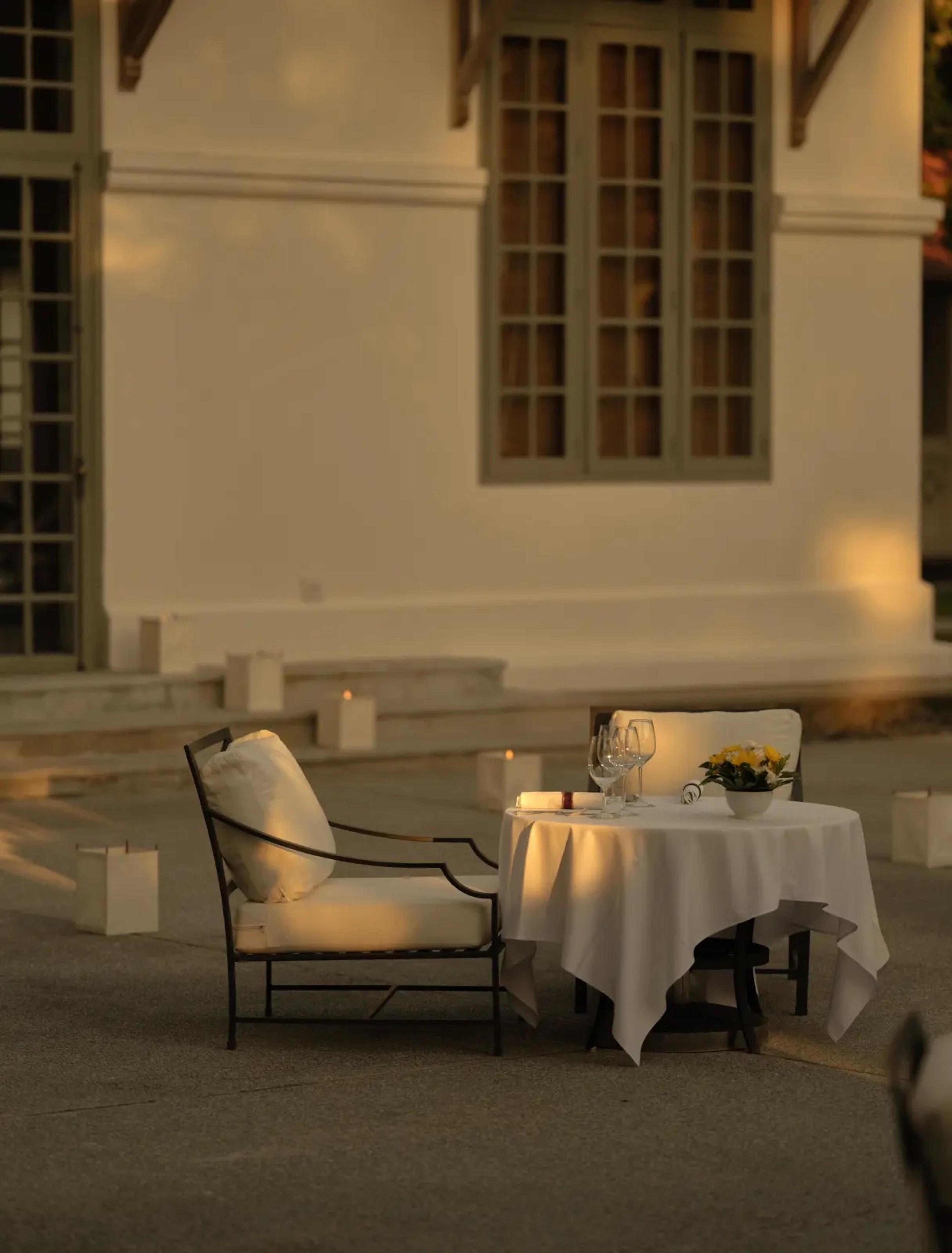 A table laid for two with wine glasses and candles beside an armchair, set against a white wall lit gold by the low sun.