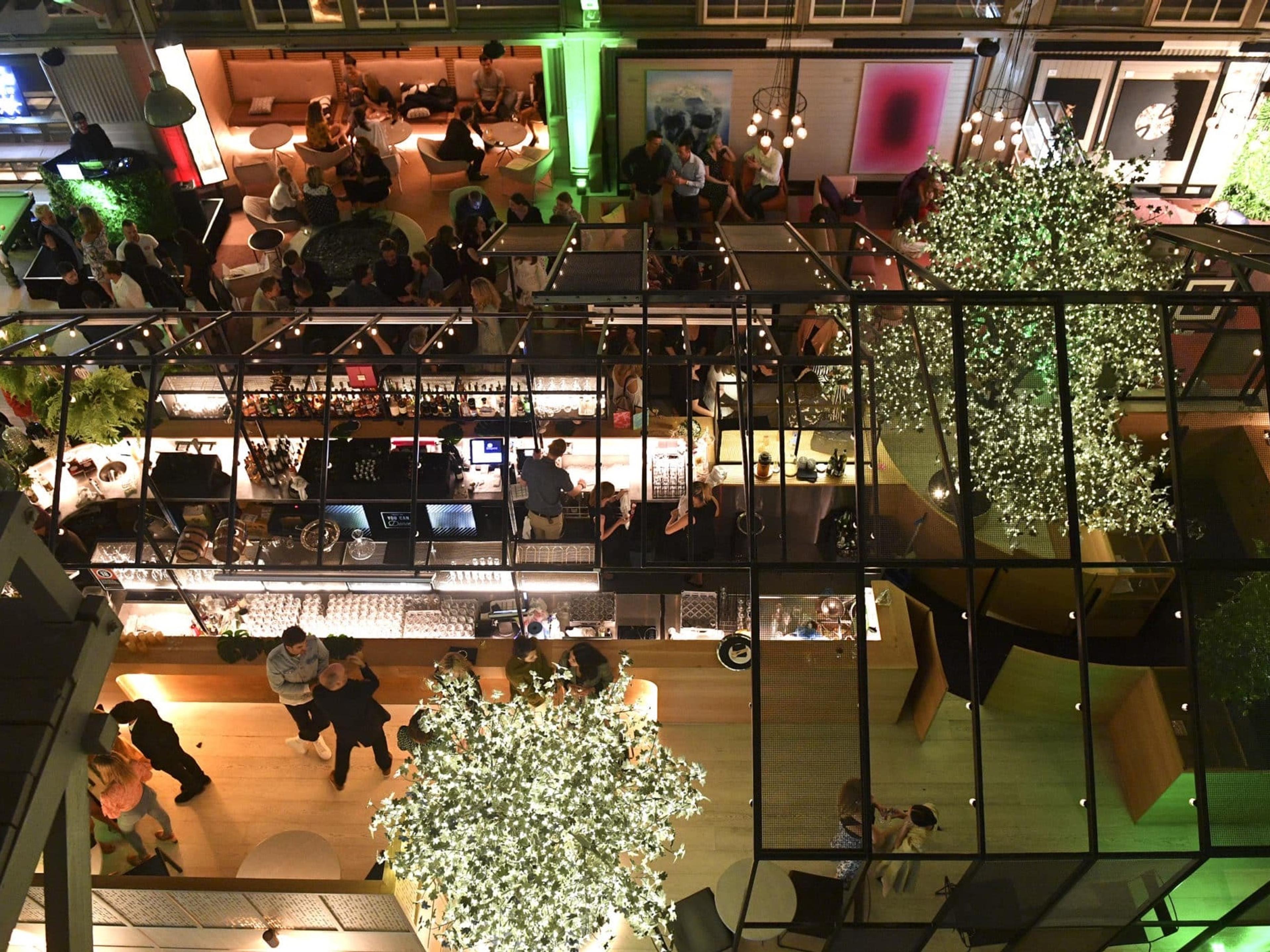 A glass-roofed courtyard bar at night seen from above, strung with lights and planted with trees