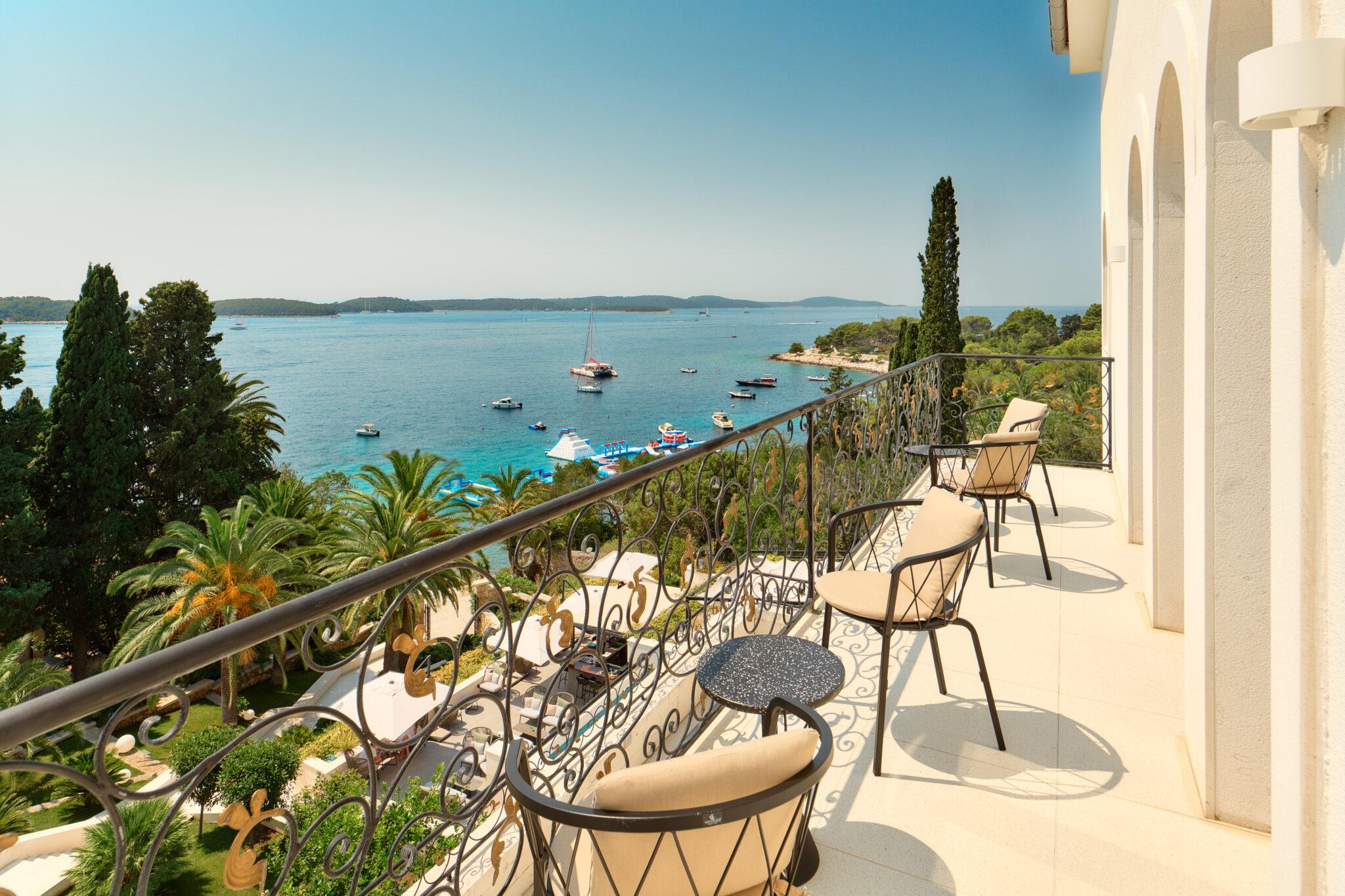 A hotel balcony with wrought iron railings and two chairs, overlooking a bay with anchored boats and yachts.