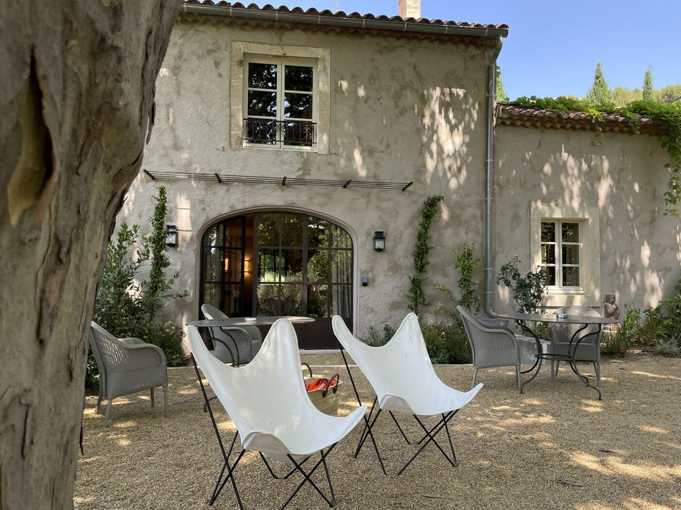 A stone farmhouse with shuttered windows and an arched doorway, with white butterfly chairs and wicker tables set on gravel in front.