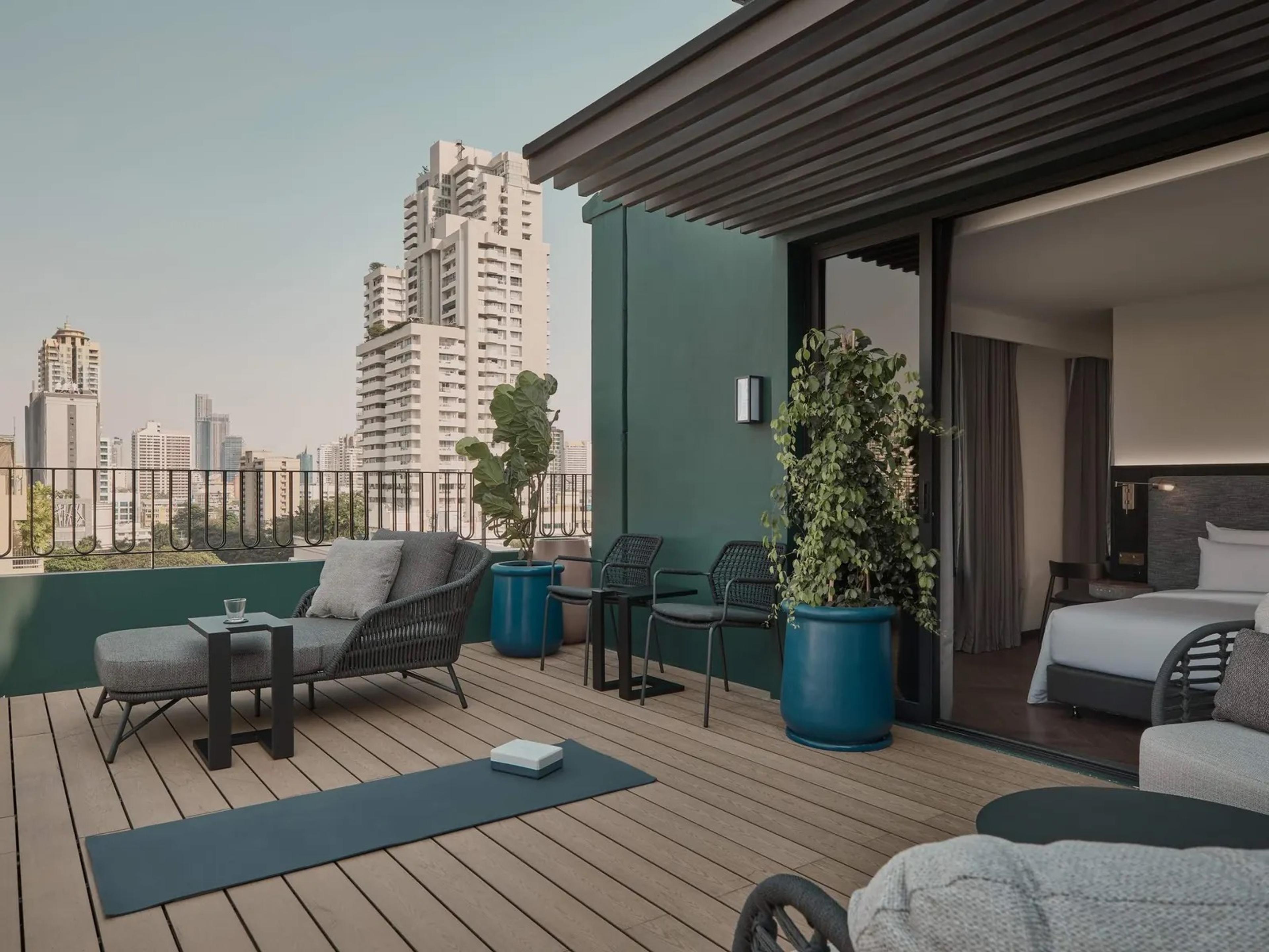 A private rooftop terrace with a sofa, chairs and potted plants, a yoga mat laid out on the decking and the city skyline beyond the railing.