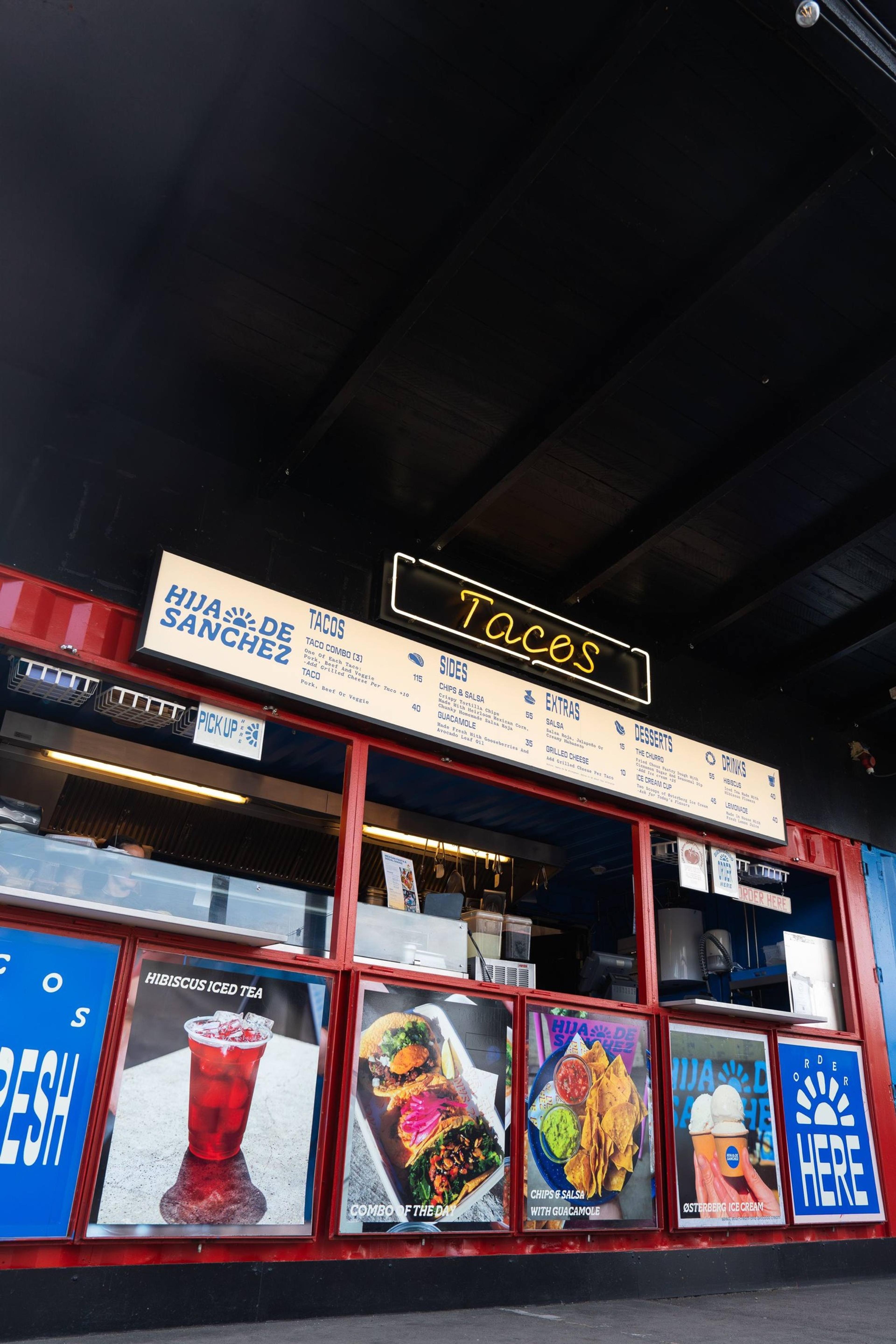 The counter at Hija de Sanchez, a red-framed hatch with the menu board above it and a yellow neon sign reading Tacos