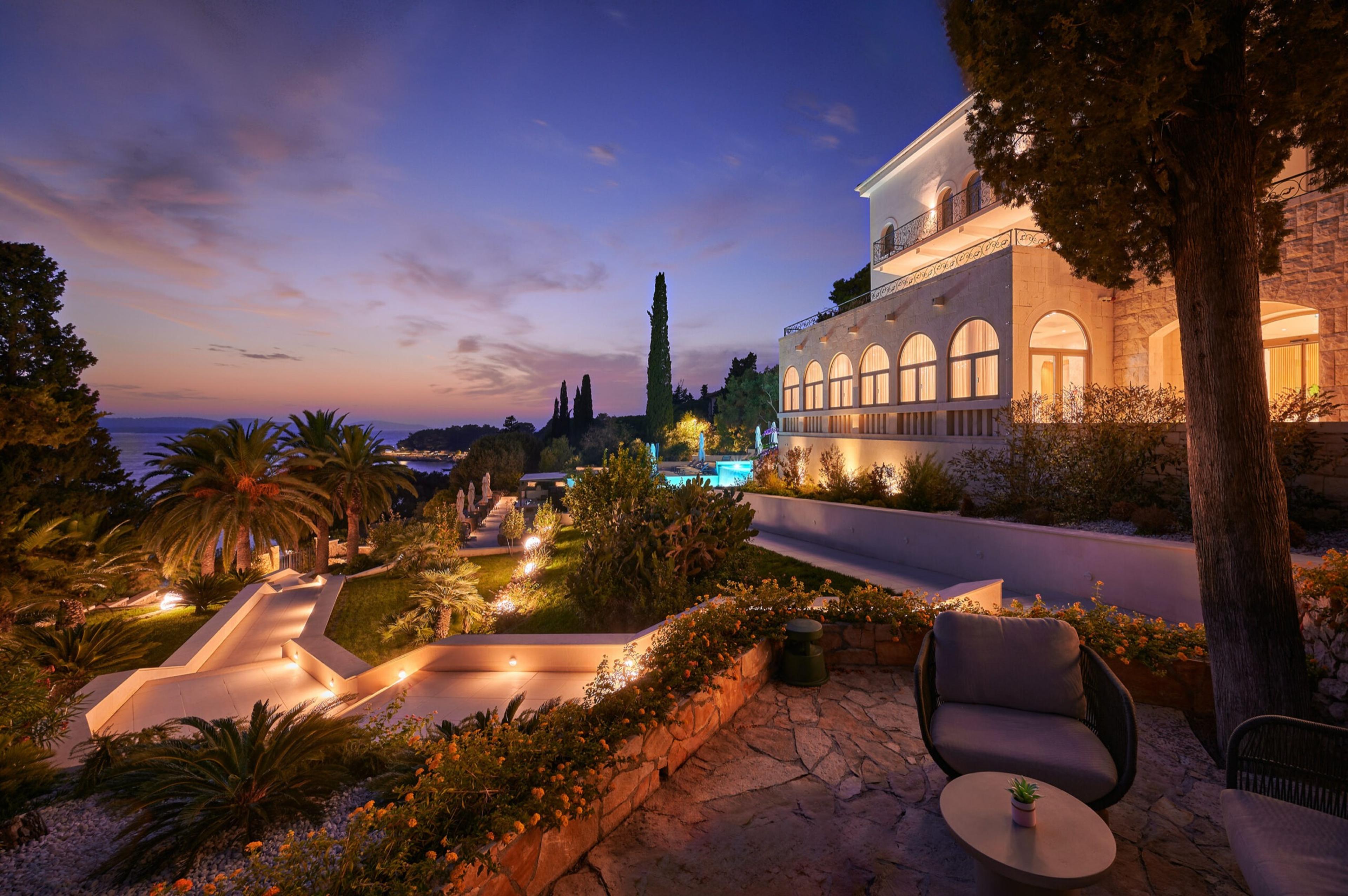 A three-storey stone villa lit up at dusk, seen across a terraced garden with palm trees, a lounge chair and a small table in the foreground.