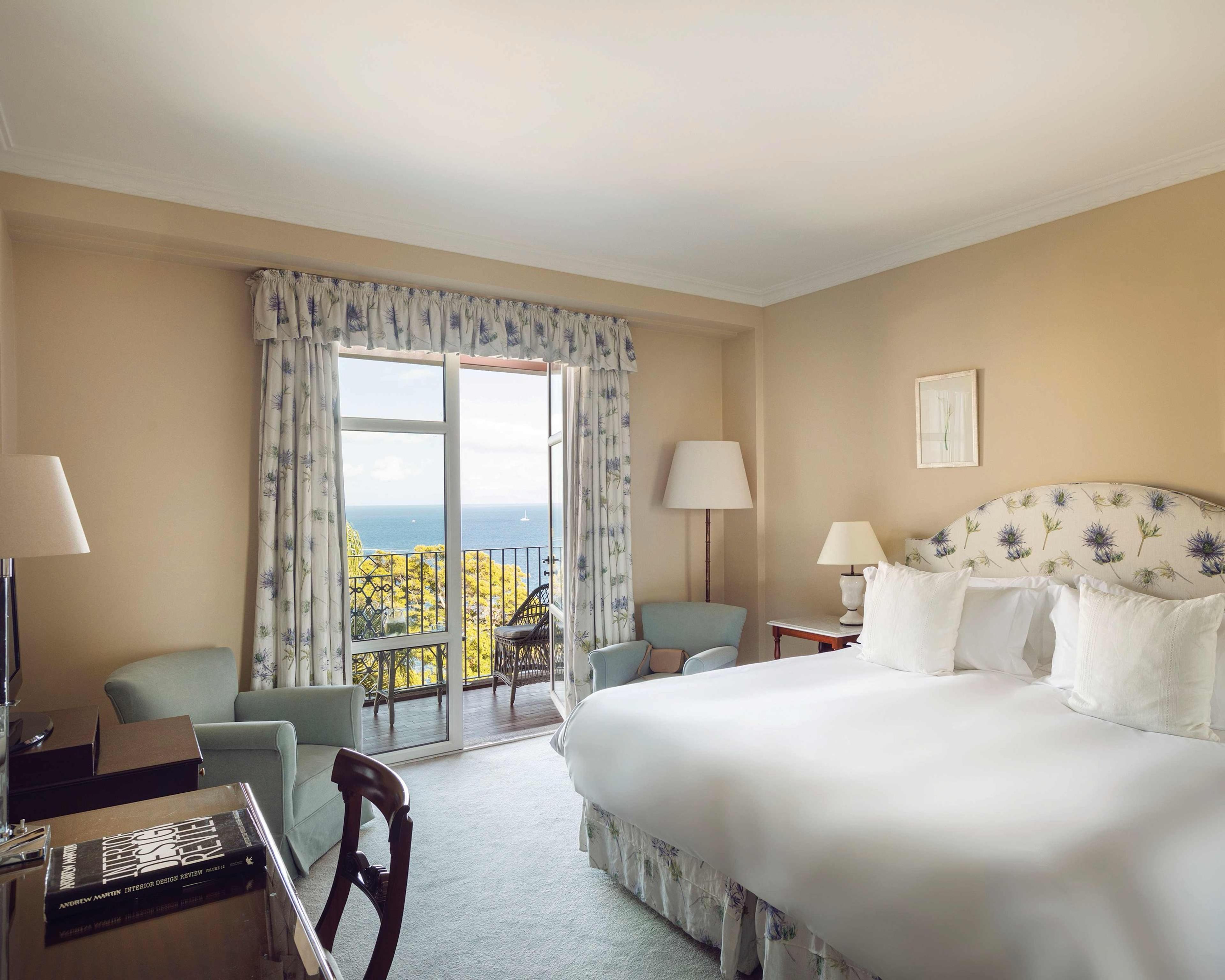 A bedroom with pale striped wallpaper has a bed with a floral headboard, two blue armchairs by the window, and a sea view through open balcony doors.