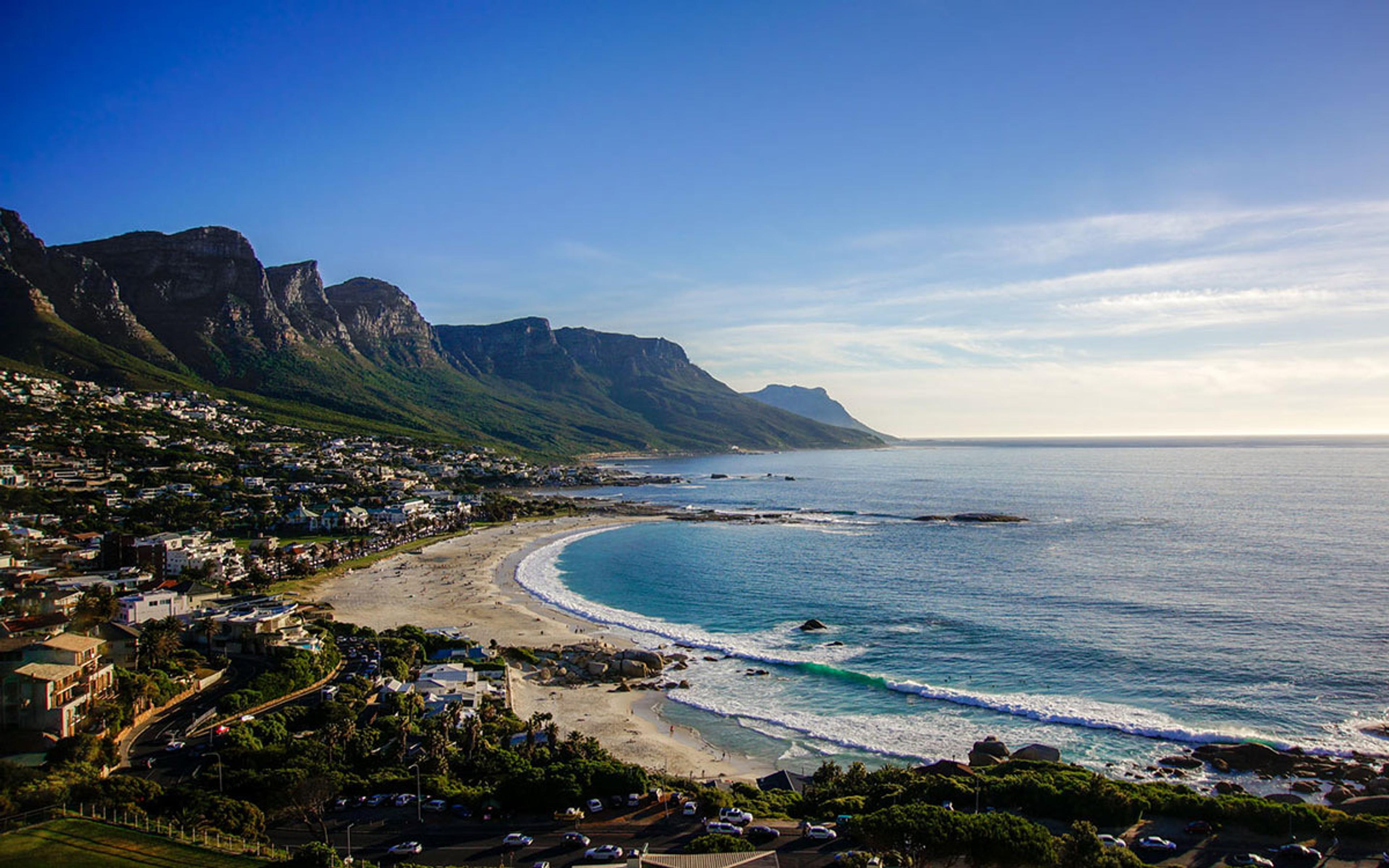 Camps Bay from above: the curved white sand and surf of the beach, the town along the left-hand shore, and a ridge of mountains rising behind the bay.