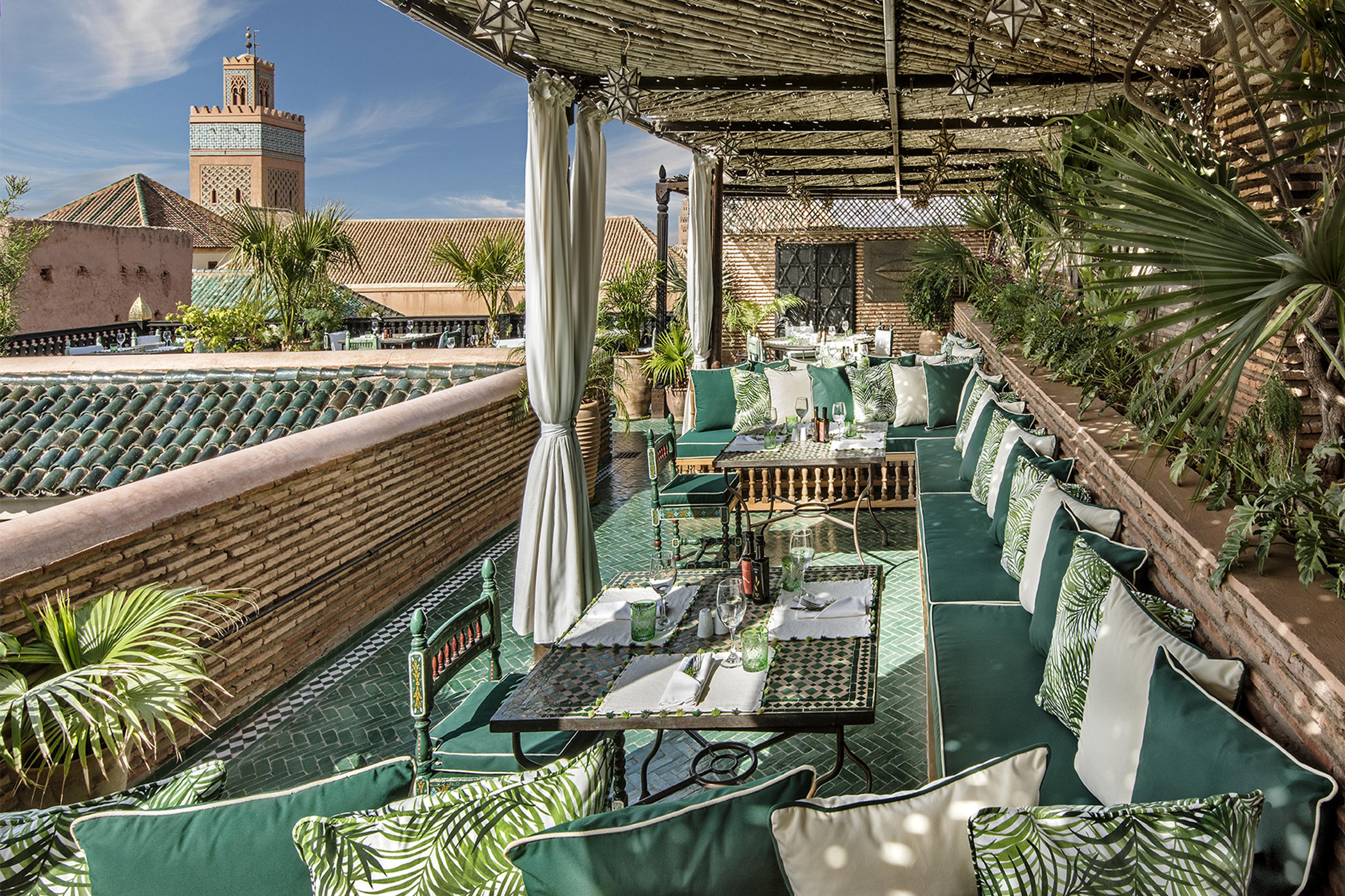 A covered rooftop terrace laid for lunch with green cushioned banquettes, looking out over tiled rooftops to a minaret under a blue sky.