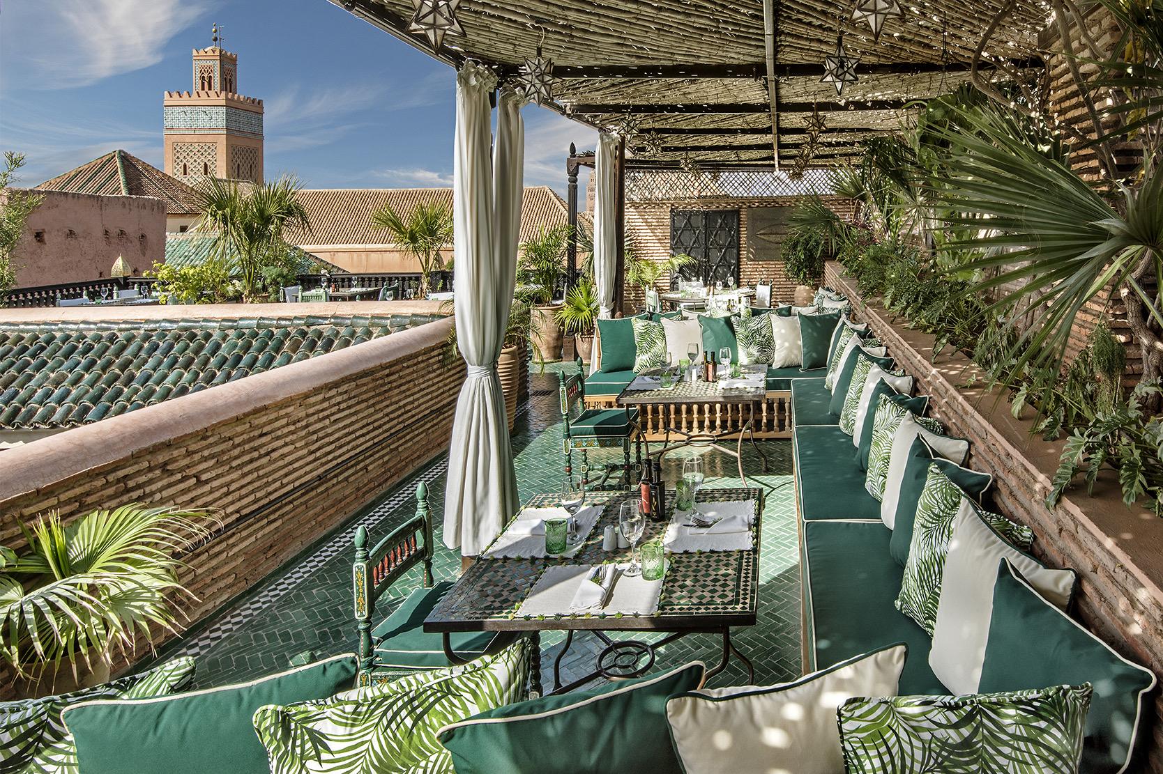 A covered rooftop terrace laid for lunch with green cushioned banquettes, looking out over tiled rooftops to a minaret under a blue sky.