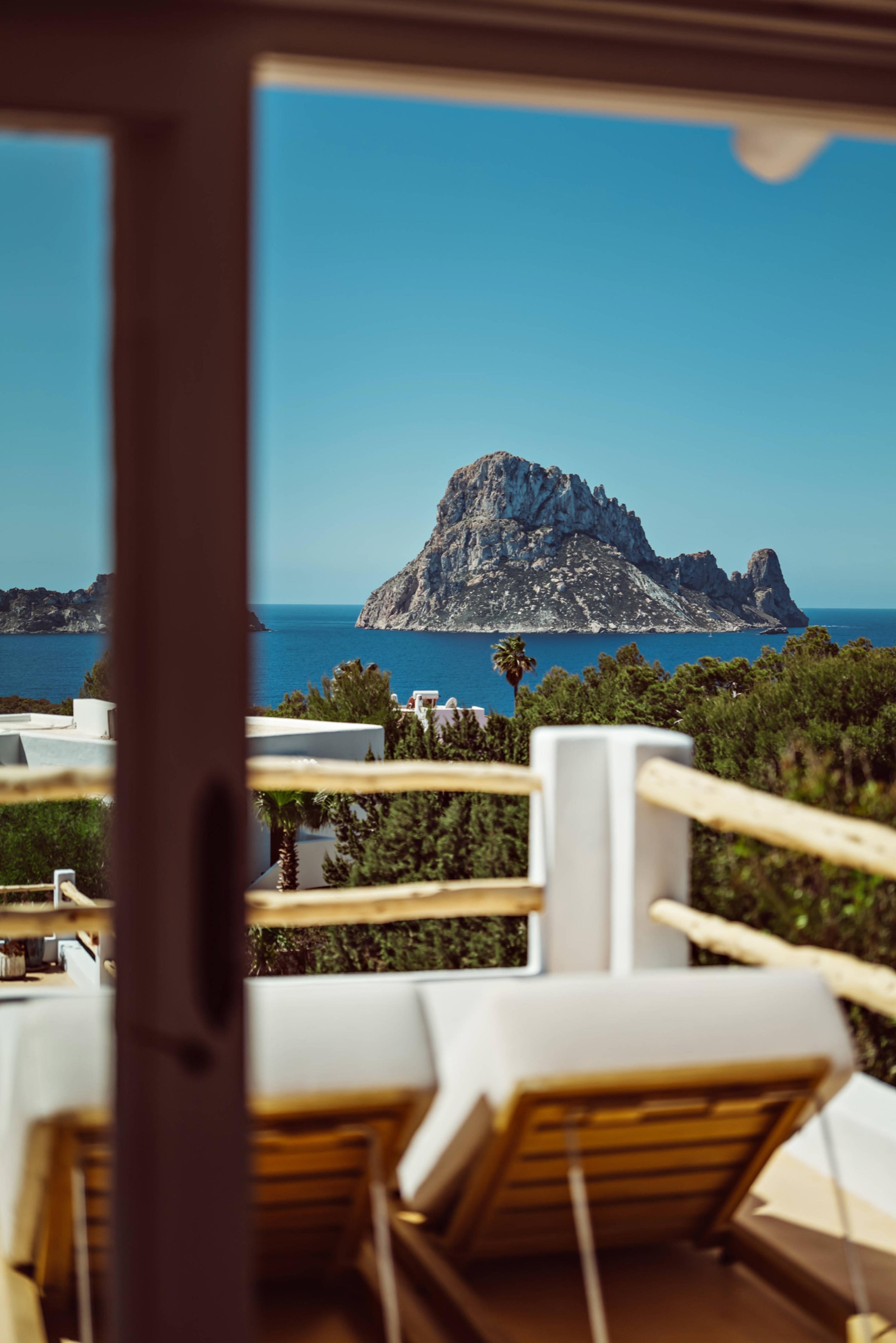 Through an open door frame, a terrace with white-cushioned sun loungers and a wooden rail, looking across pine trees to a rocky island in a blue sea.