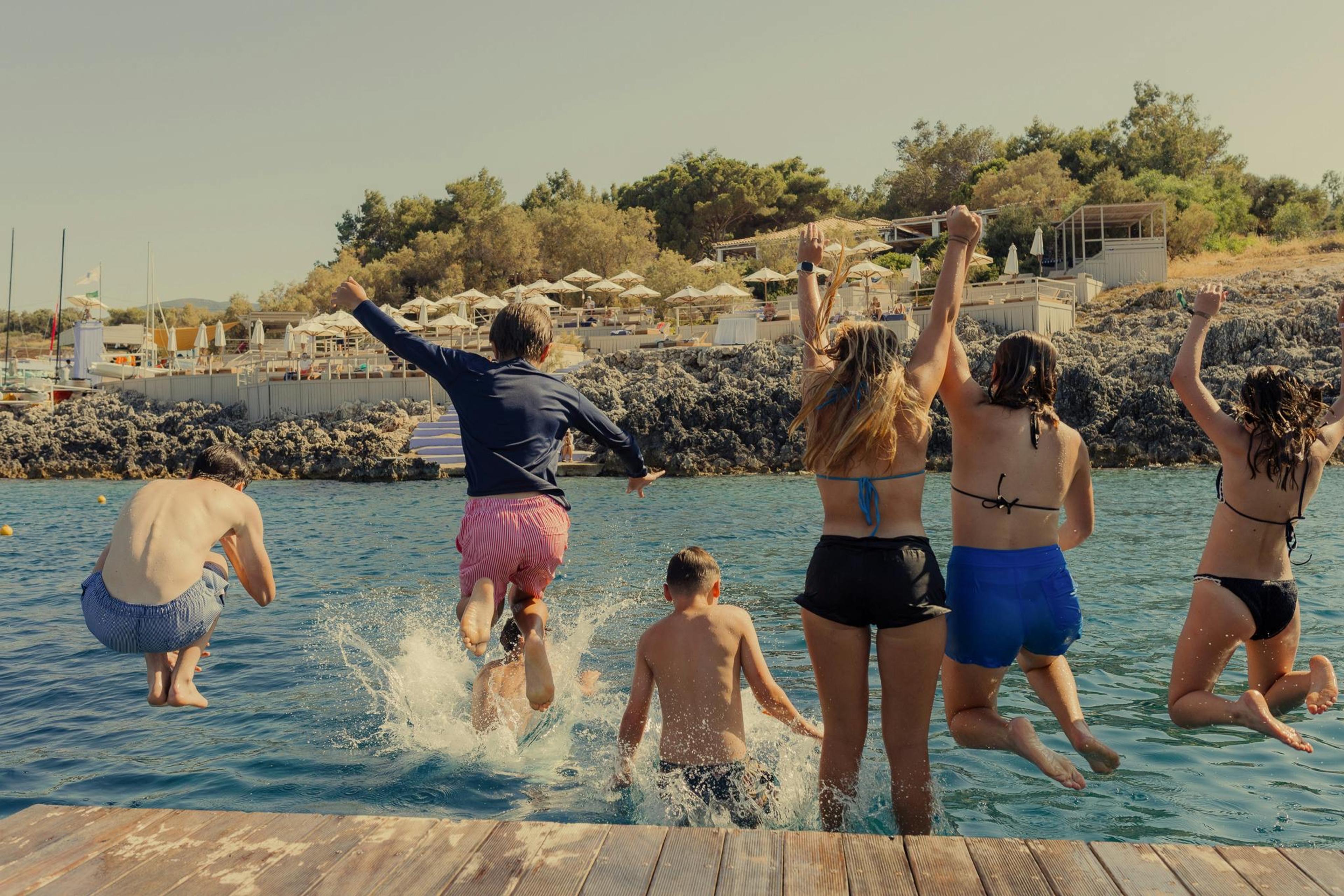 Five teenagers and children jump together off a wooden dock into the sea, a beach club and hillside visible behind them.
