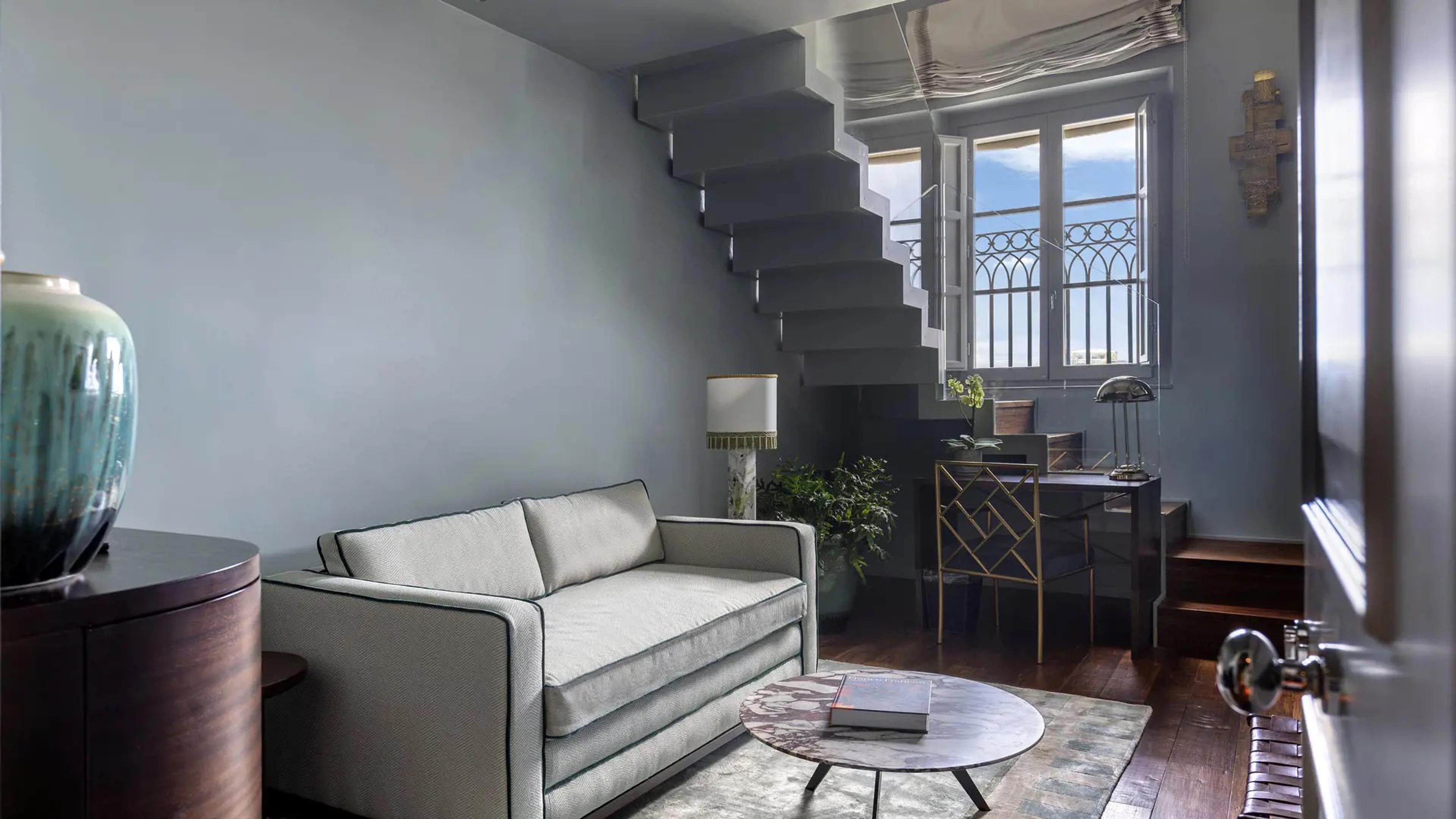 A living room with a cantilevered staircase, a pale sofa, a brass and glass desk and chair, and a window with wrought iron balcony railings.