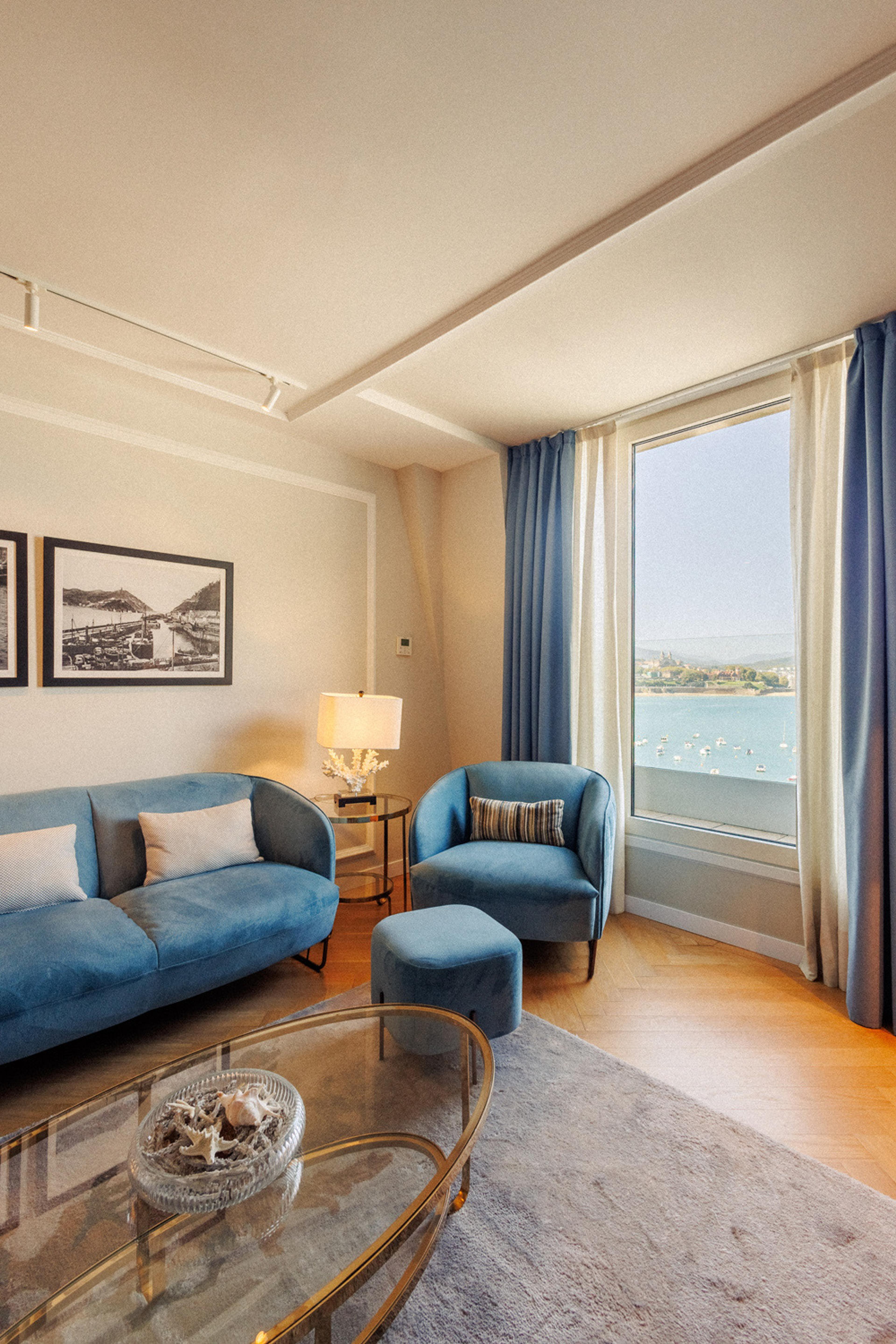 A living room with a blue sofa and armchair, black and white harbour prints on the wall, and a window over the bay.