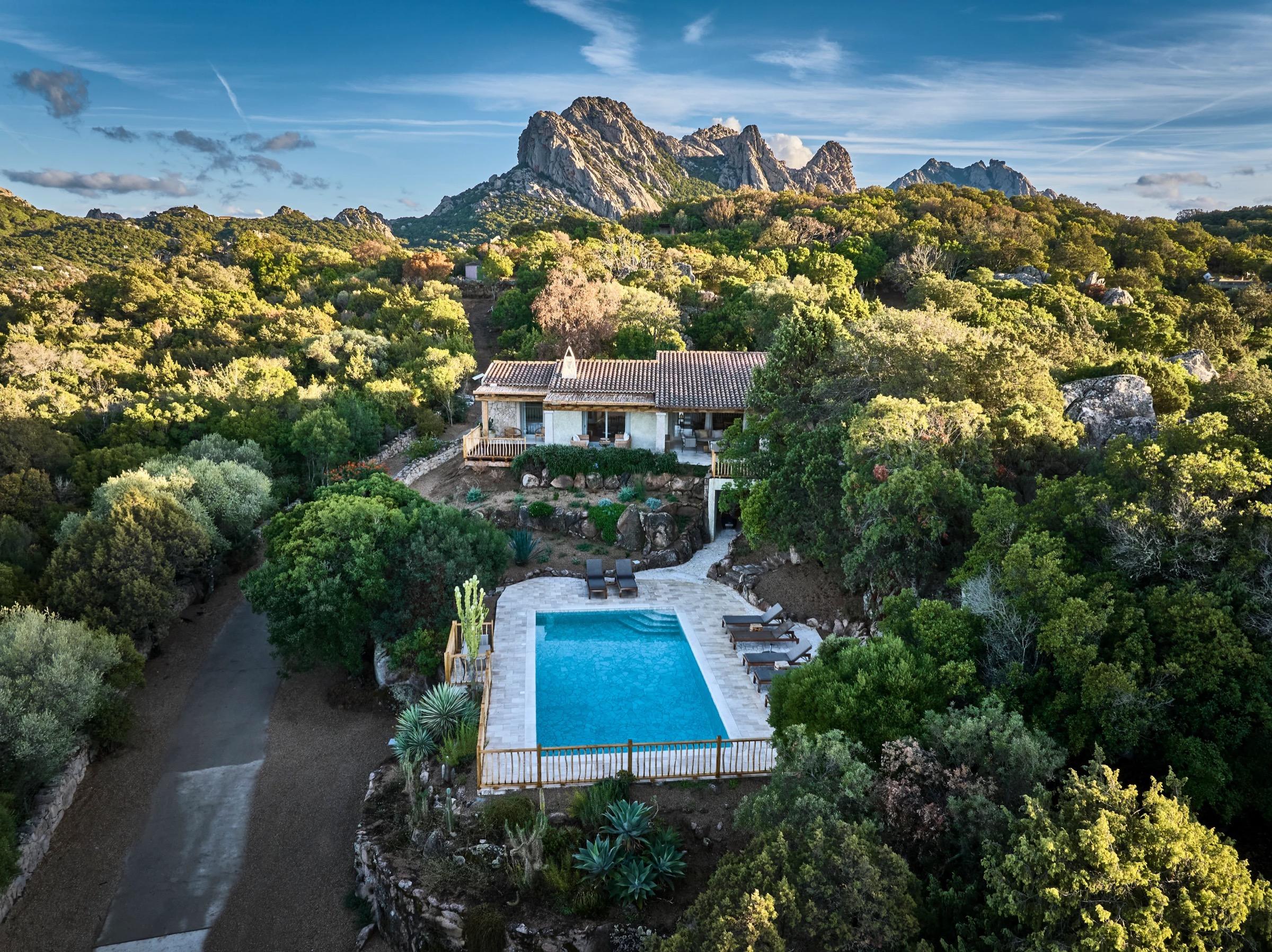 an aerial view at golden hour of a stone house with a tiled roof and a swimming pool, surrounded by trees with rocky peaks behind