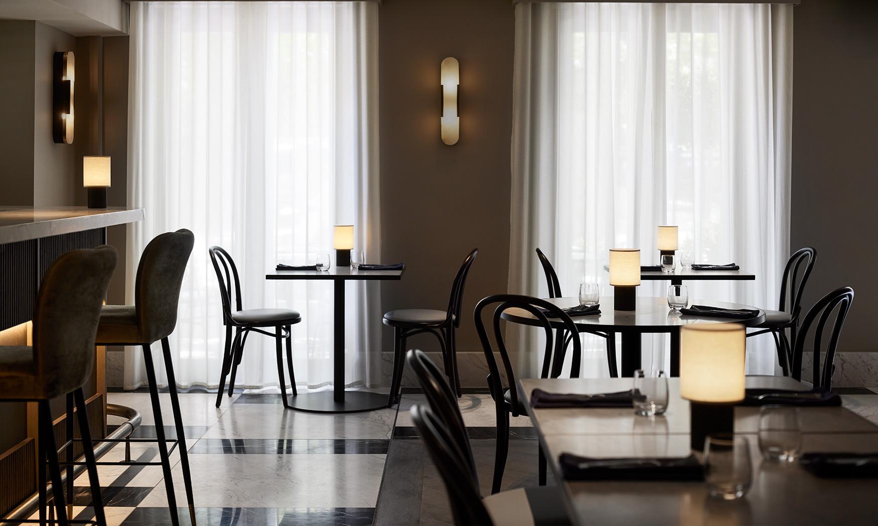A dining room with marble floors, bentwood chairs and tables laid for service beside tall curtained windows.