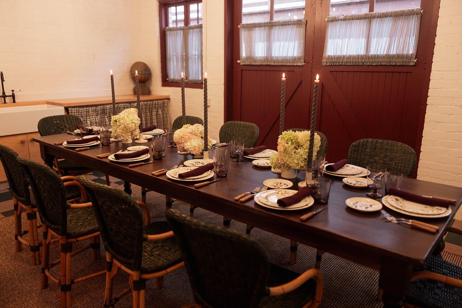 A long wooden table laid with patterned plates, dark napkins, lit taper candles and hydrangea arrangements, in front of two dark red barn doors.