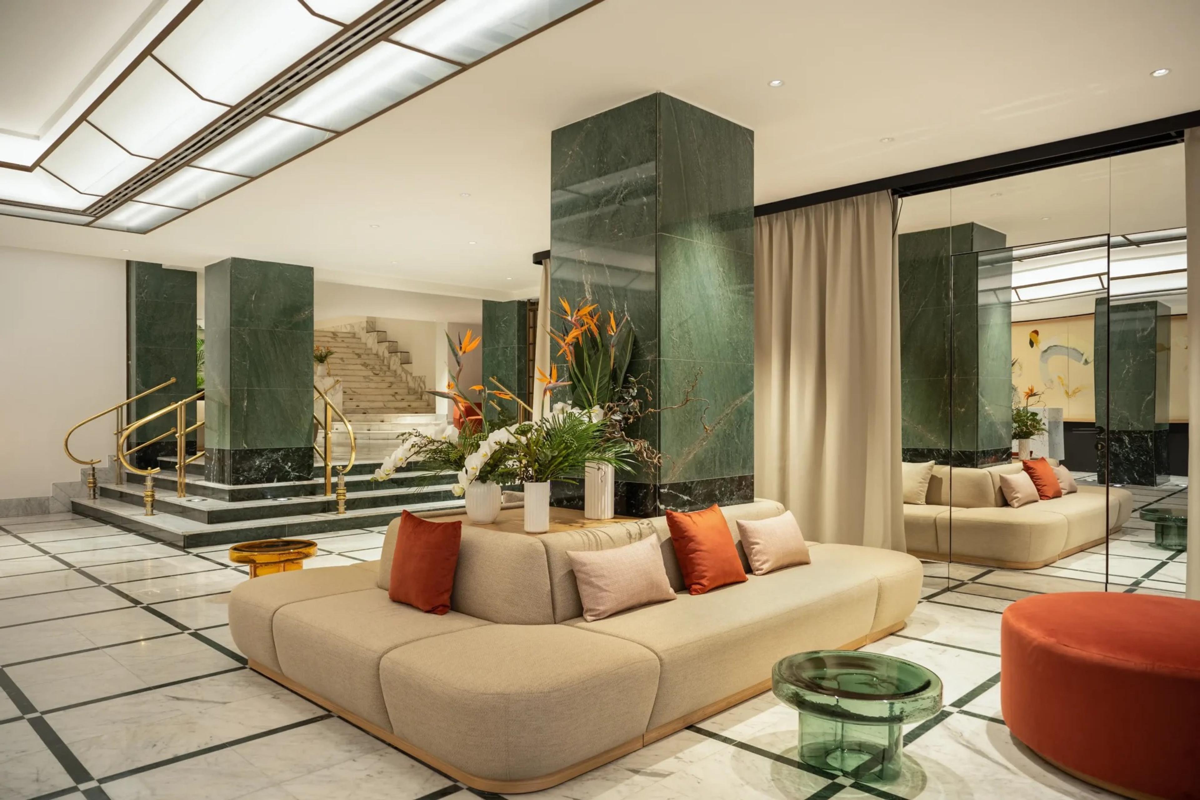 A lobby with green marble columns, a cream curved sofa with orange and pink cushions, white orchids in vases, and a staircase behind brass rails.
