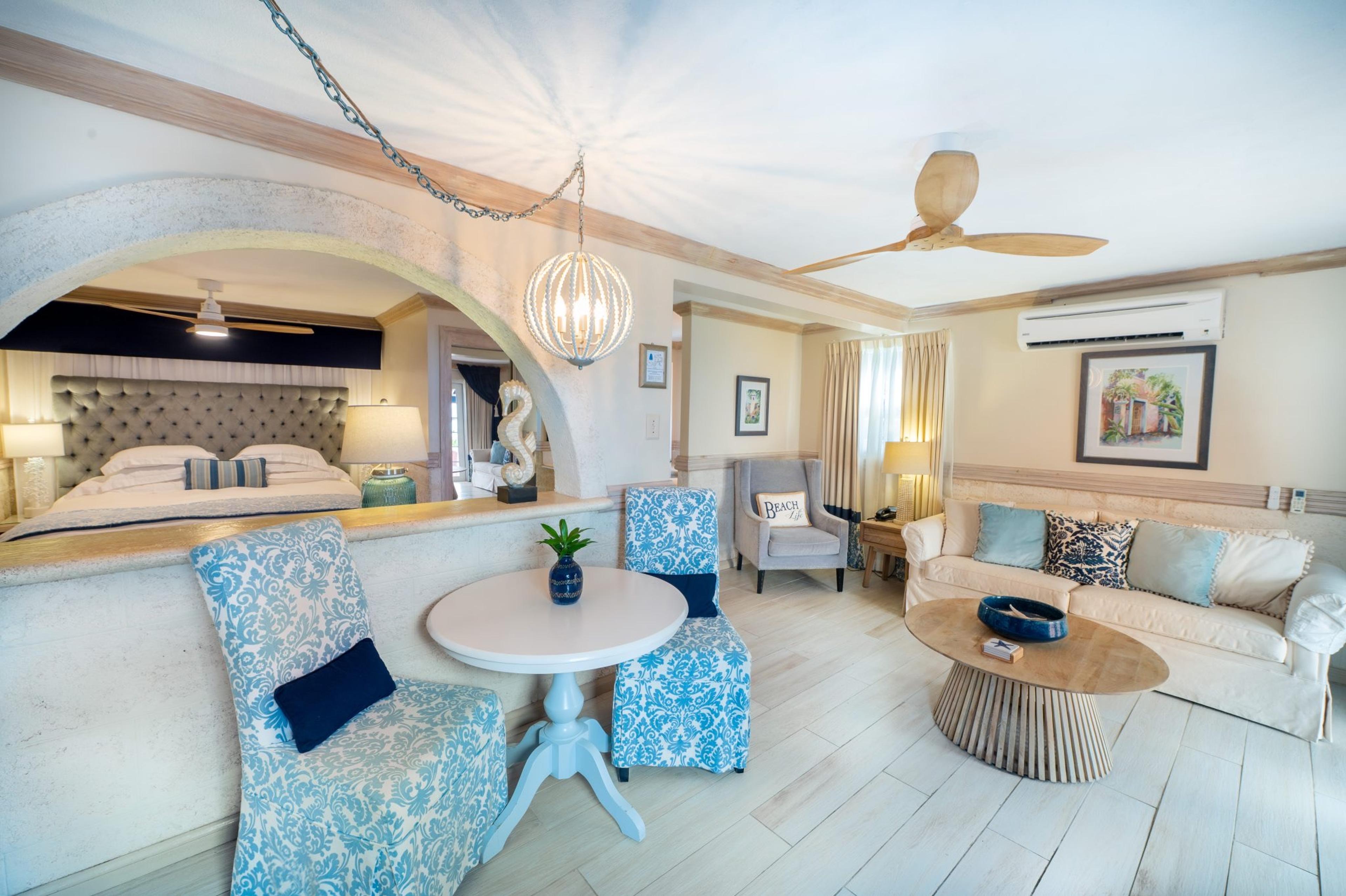 Living area of a hotel suite with a round table flanked by blue patterned chairs, and an archway opening onto a bed with a grey tufted headboard.