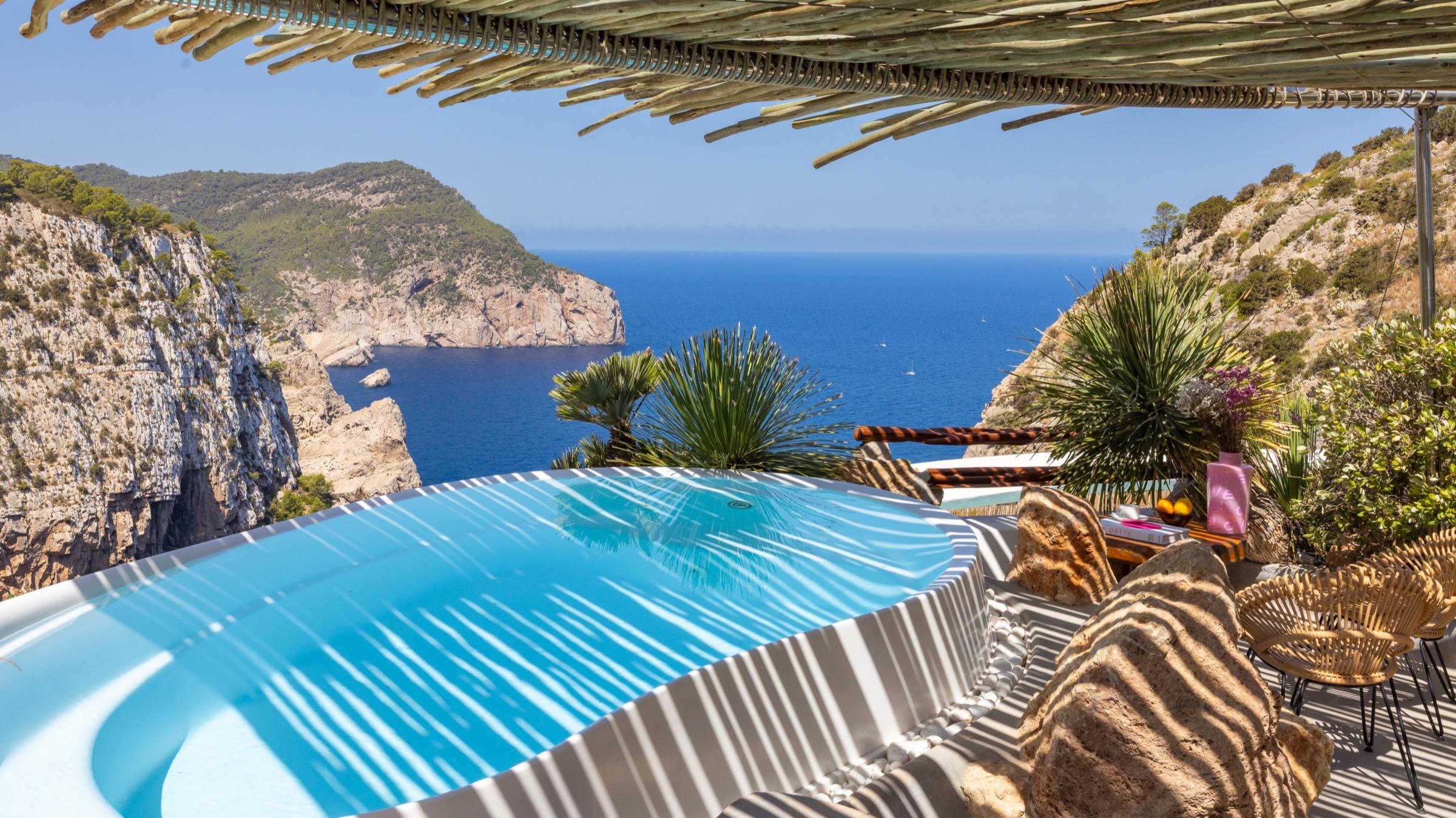 A round pool under a reed canopy on a clifftop, with rocky headlands, pines and the open sea beyond.