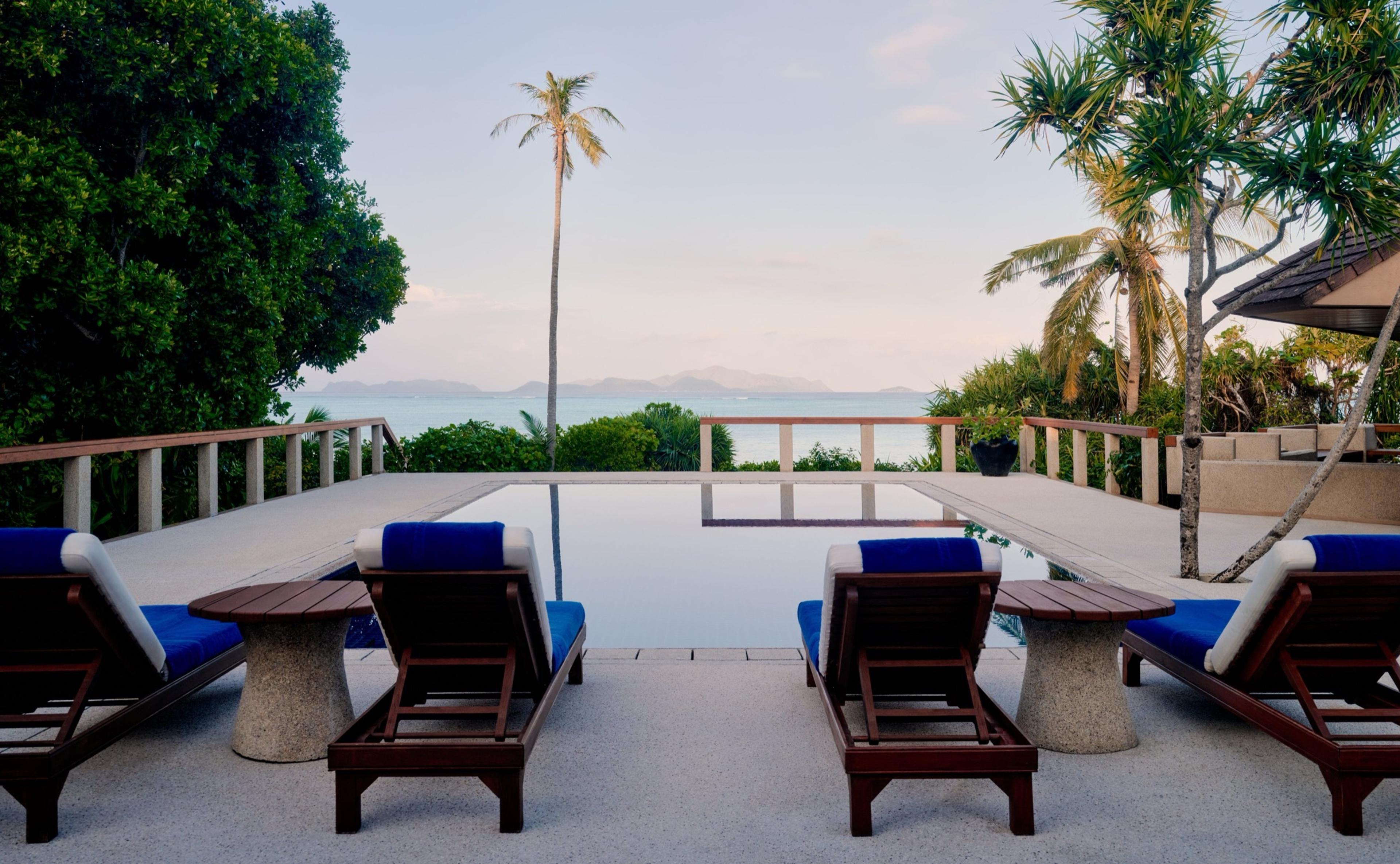 Four lounge chairs face an infinity pool at dusk, with the sea and distant hills beyond.