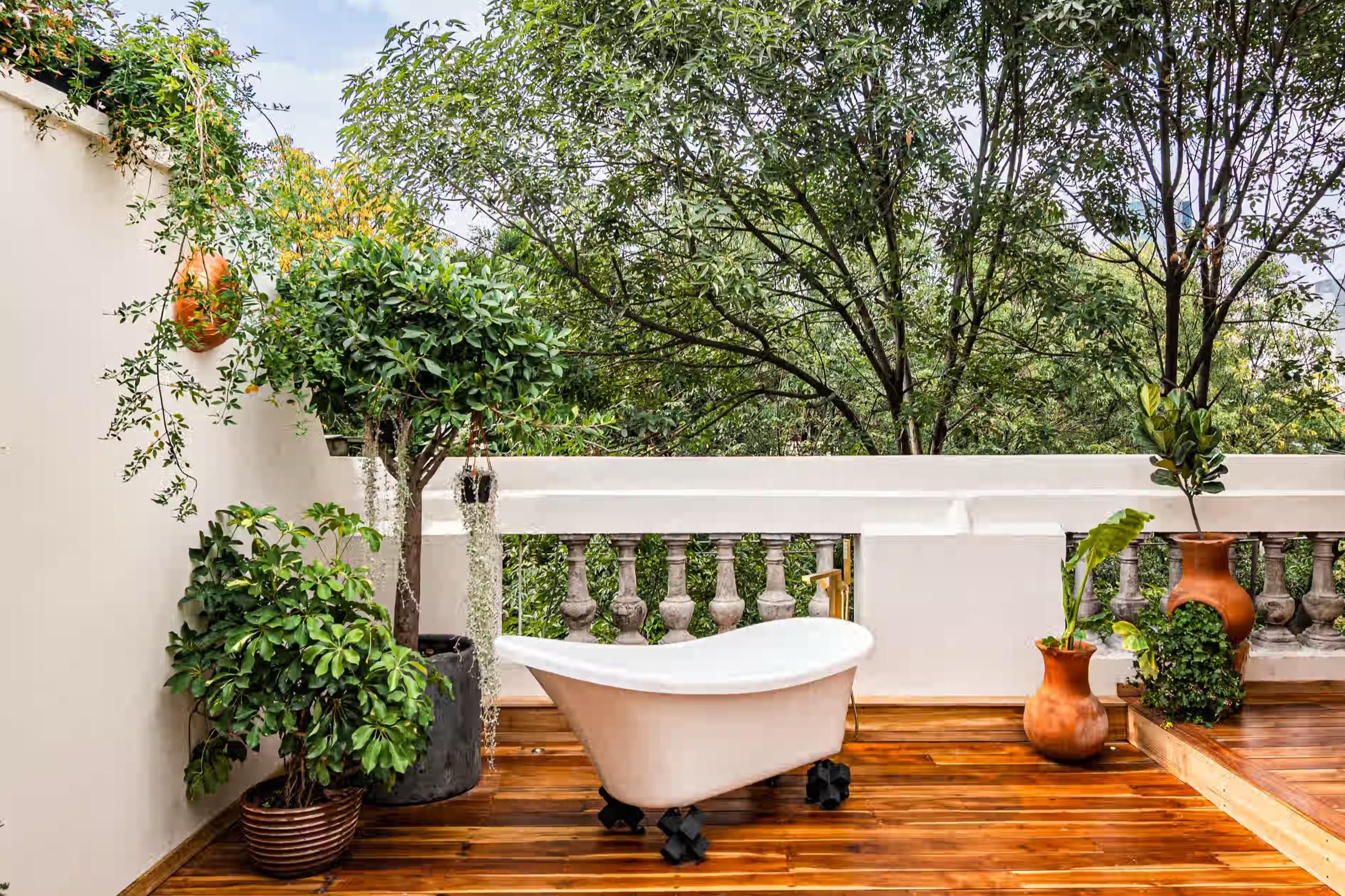 A white clawfoot bathtub stands on a wooden deck among potted trees and a stone balustrade.