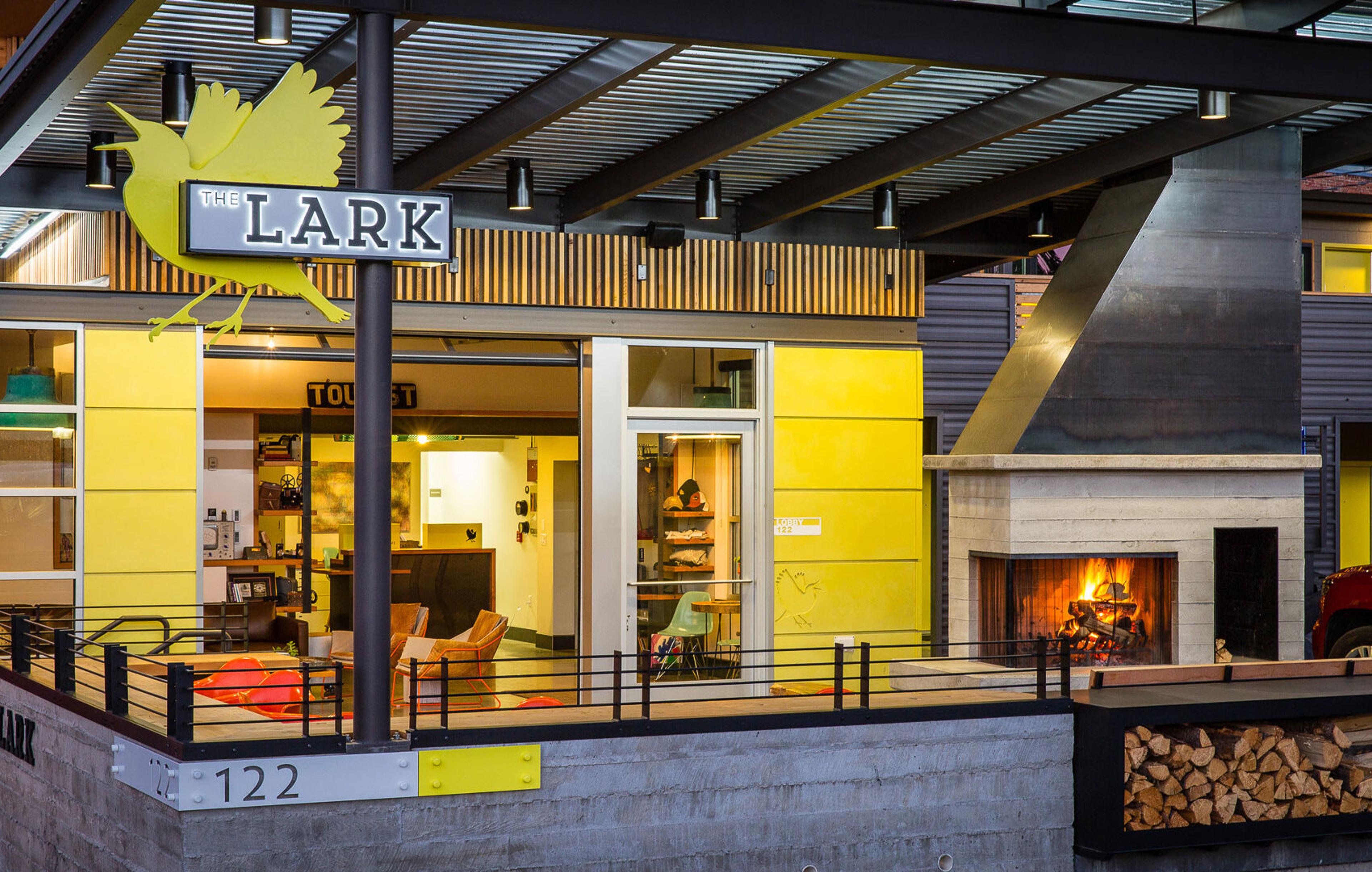 The front of a hotel at dusk under a covered roof, with a yellow bird sign reading THE LARK above the entrance and a lit fireplace on the outdoor terrace.