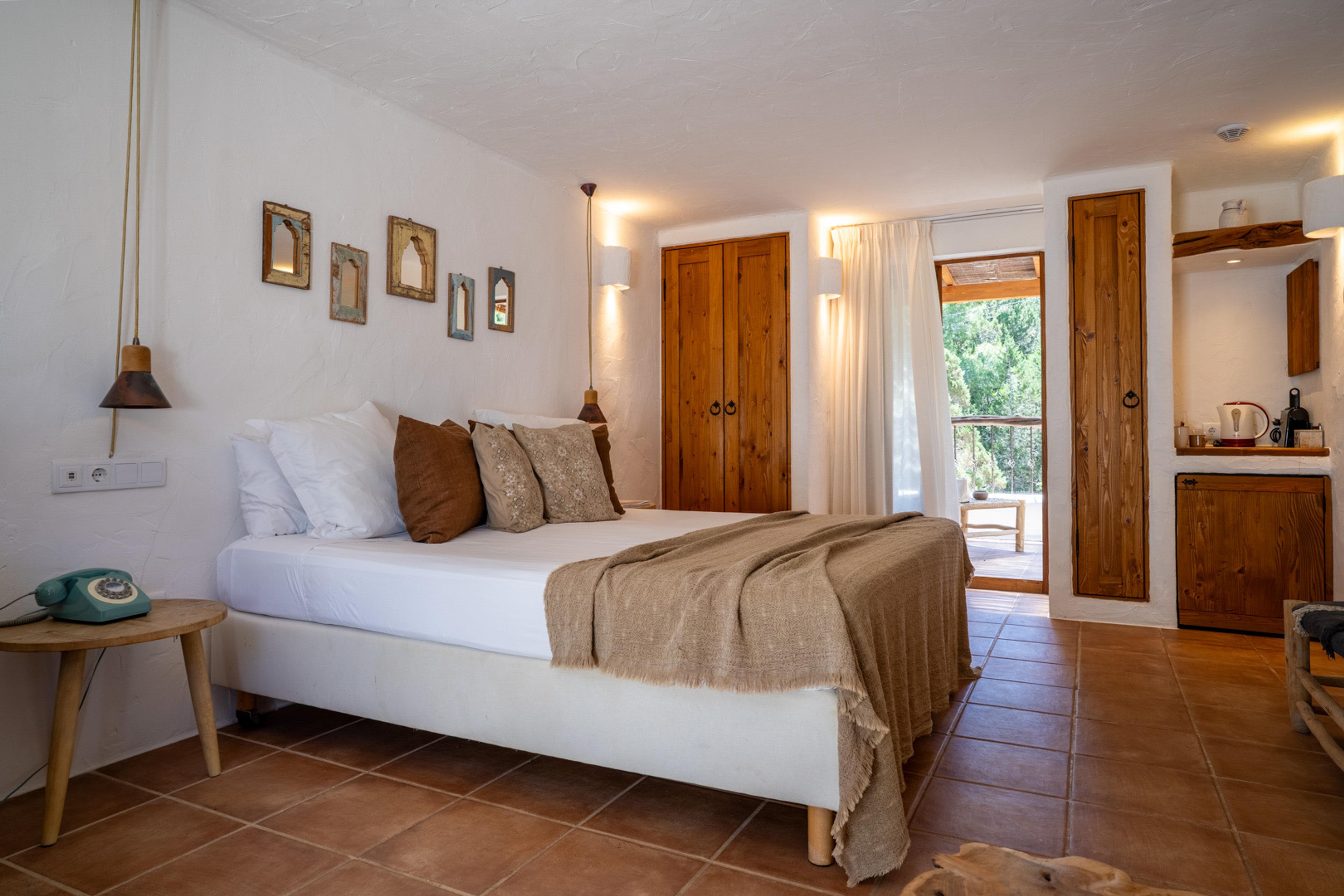 An open-plan bedroom with a white bed and patterned pillows, wooden wardrobe doors, and a kitchenette visible through the doorway.
