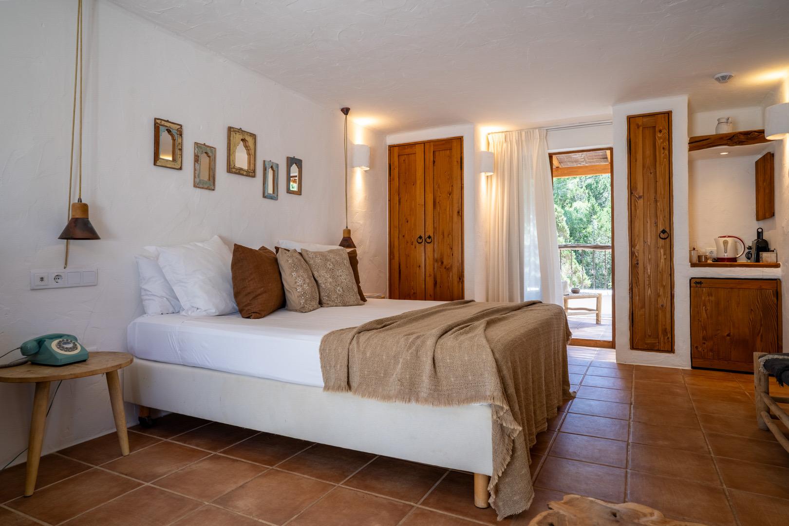 An open-plan bedroom with a white bed and patterned pillows, wooden wardrobe doors, and a kitchenette visible through the doorway.