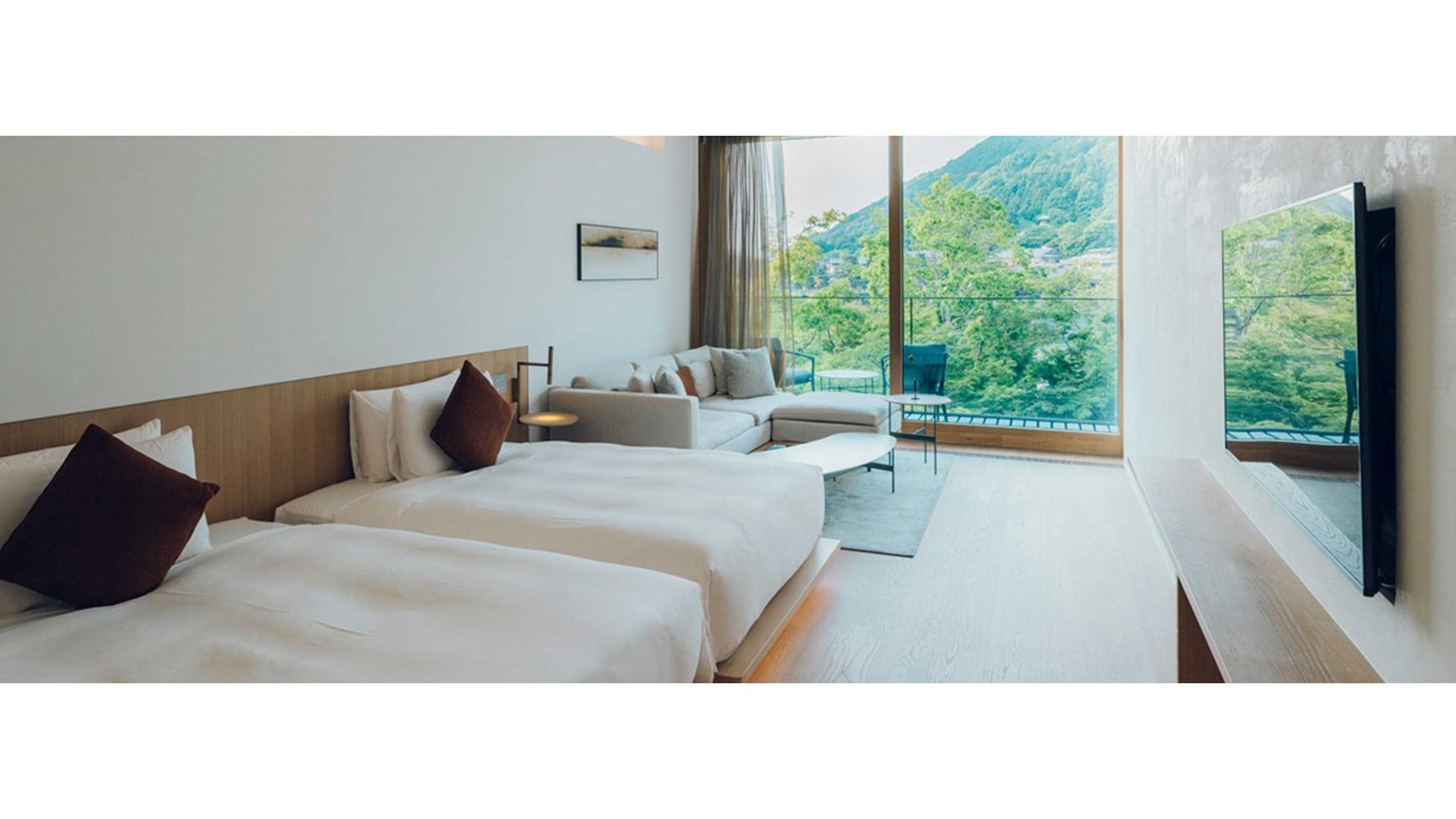 A hotel bedroom with two beds and a corner sofa, a full-height window showing a wooded mountainside and balcony