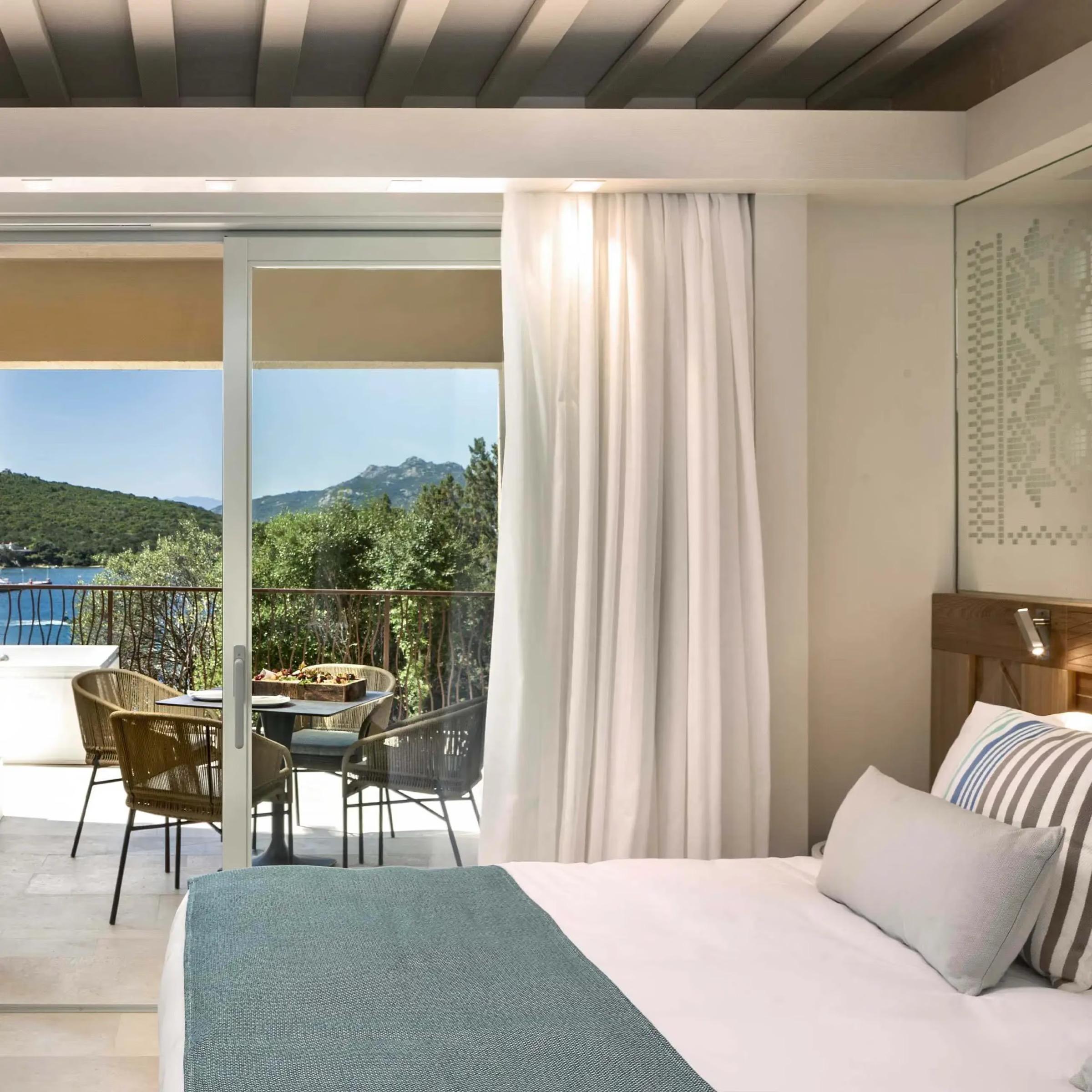 A bedroom with a patterned headboard opens through glass doors onto a terrace with chairs overlooking a bay and wooded hills.