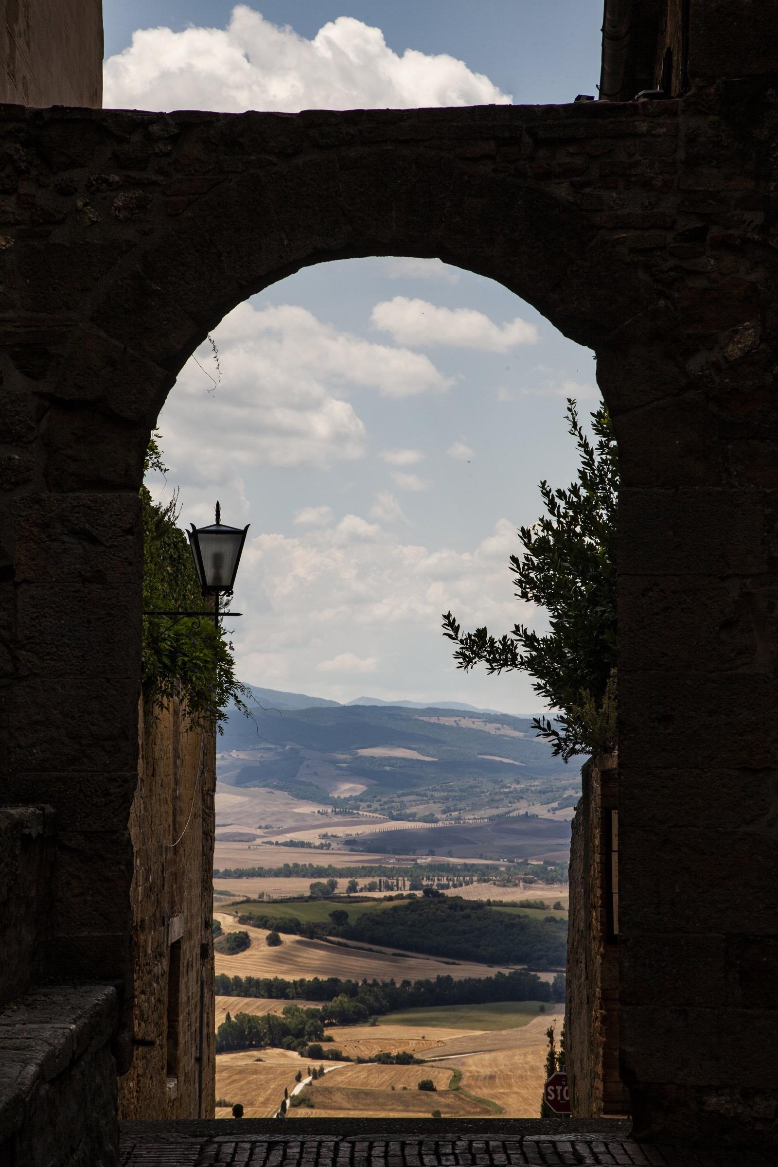 A stone archway framing a view of Tuscan farmland and hills under a partly clouded sky.
