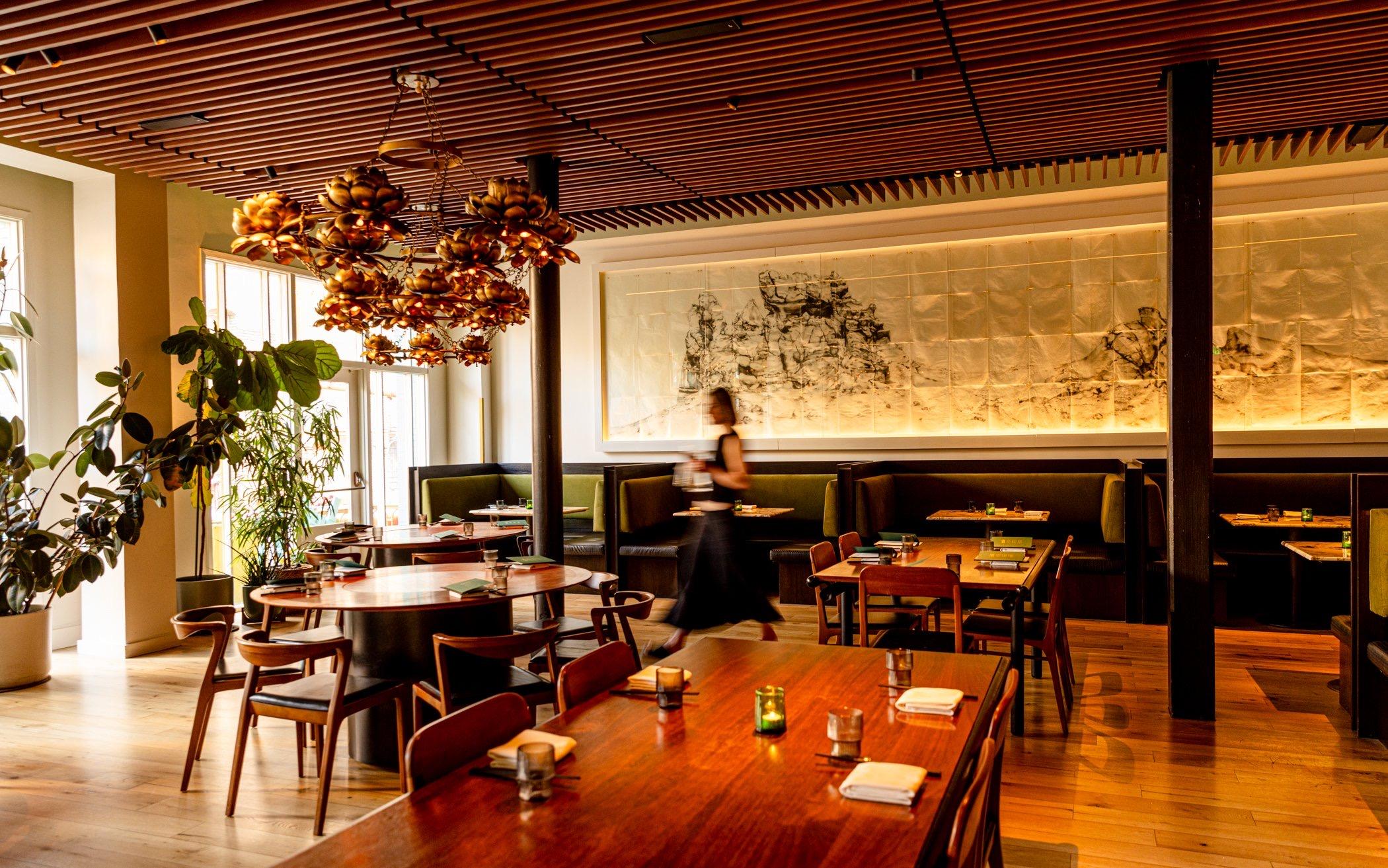 A warm wood-ceilinged dining room with brass lotus chandeliers, set tables and a long banquette under a painted mural