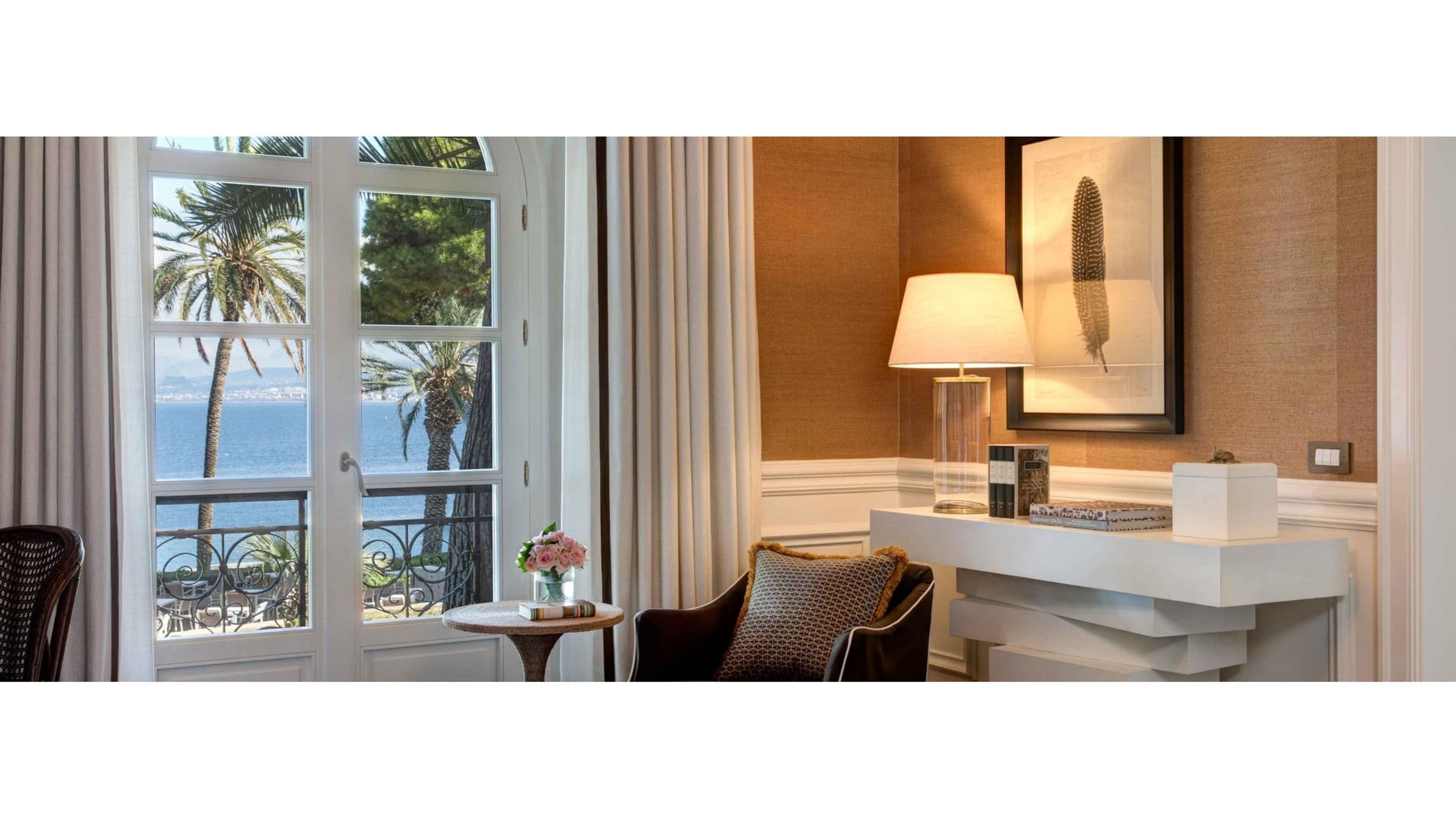 A sitting area beside open French doors onto a balcony, with palm trees and the sea visible beyond, and a small table with flowers and books by an armchair.