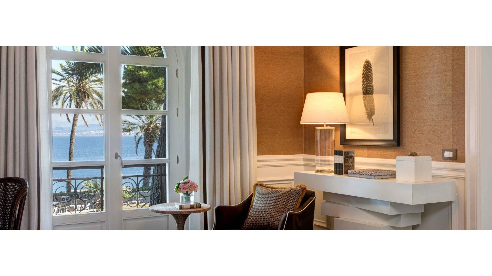 A sitting area beside open French doors onto a balcony, with palm trees and the sea visible beyond, and a small table with flowers and books by an armchair.