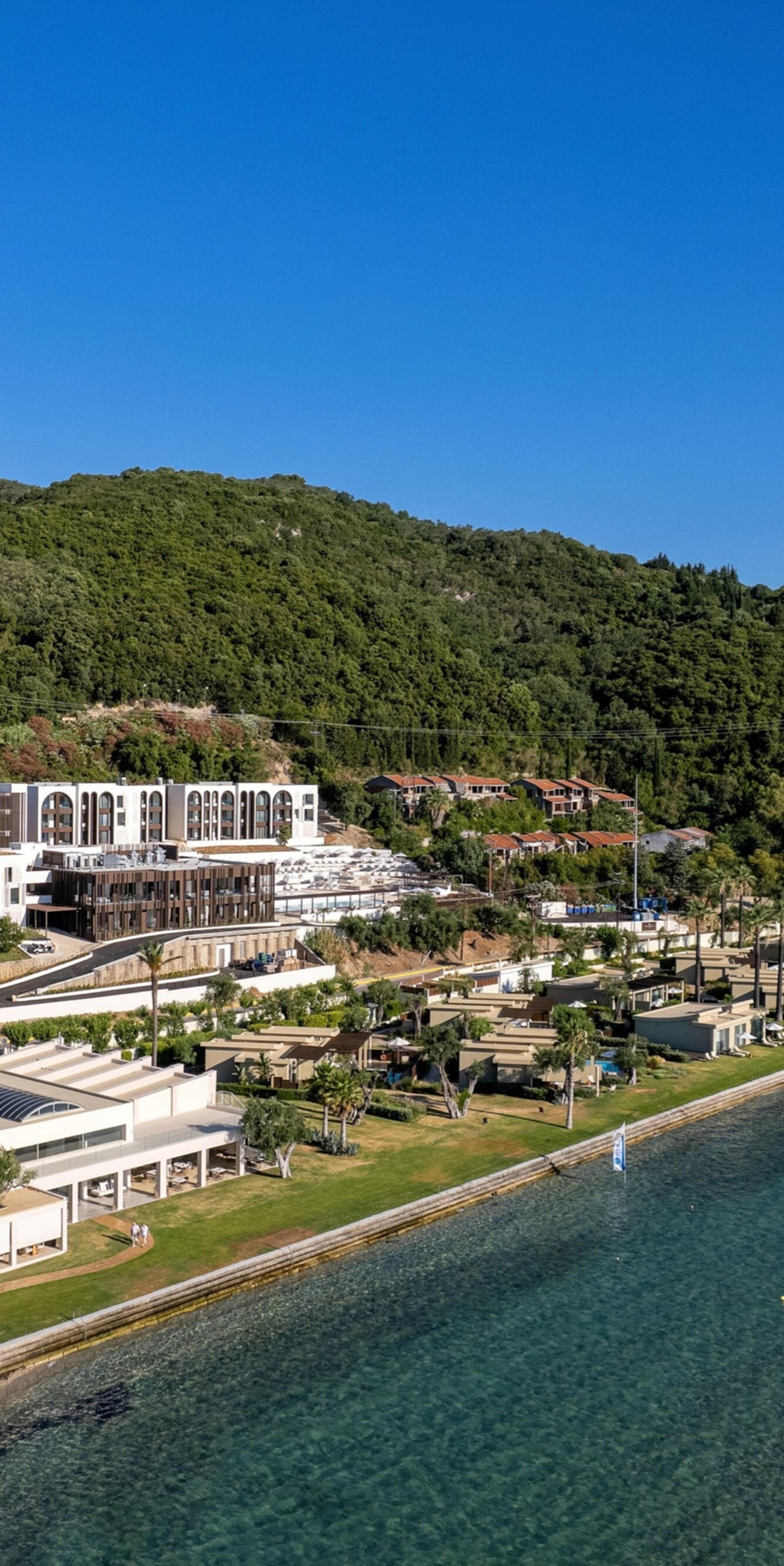 An aerial view of a resort's white buildings and low villas set among lawns and trees at the foot of a wooded hillside, with the sea in the foreground.